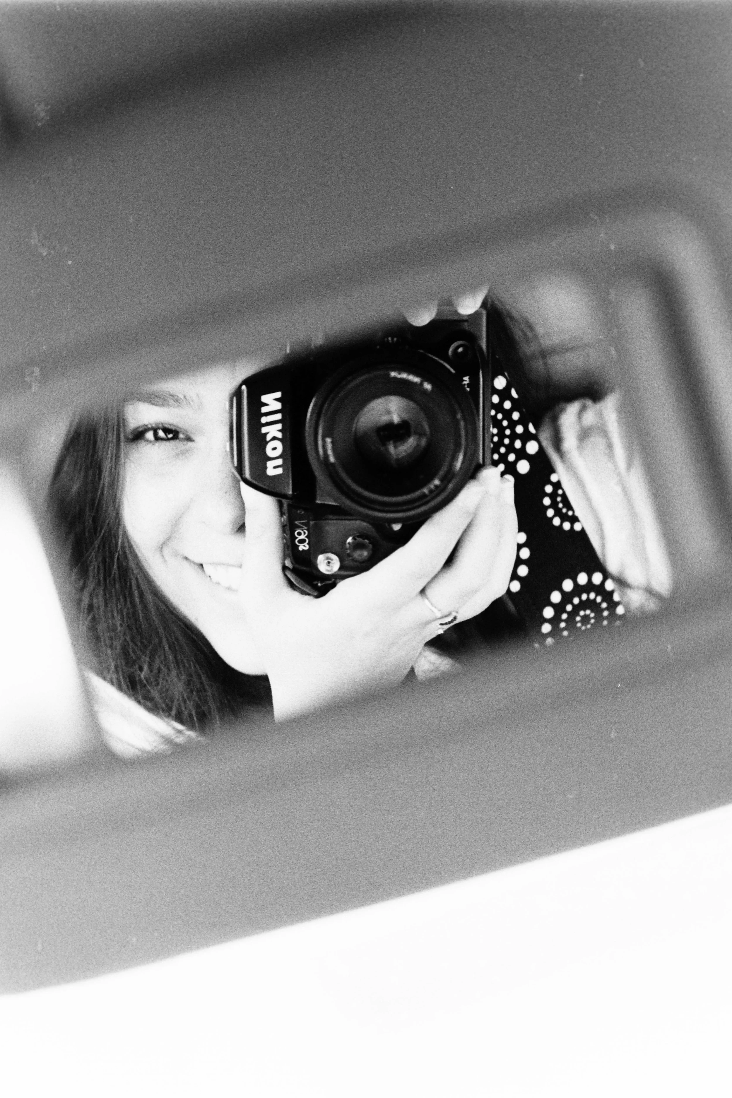 A person with light skin and long hair taking a photo of themselves with a digital camera in a mirror. The person is smiling, winking, and wearing a patterned shirt, reflected in a black-and-white photo.