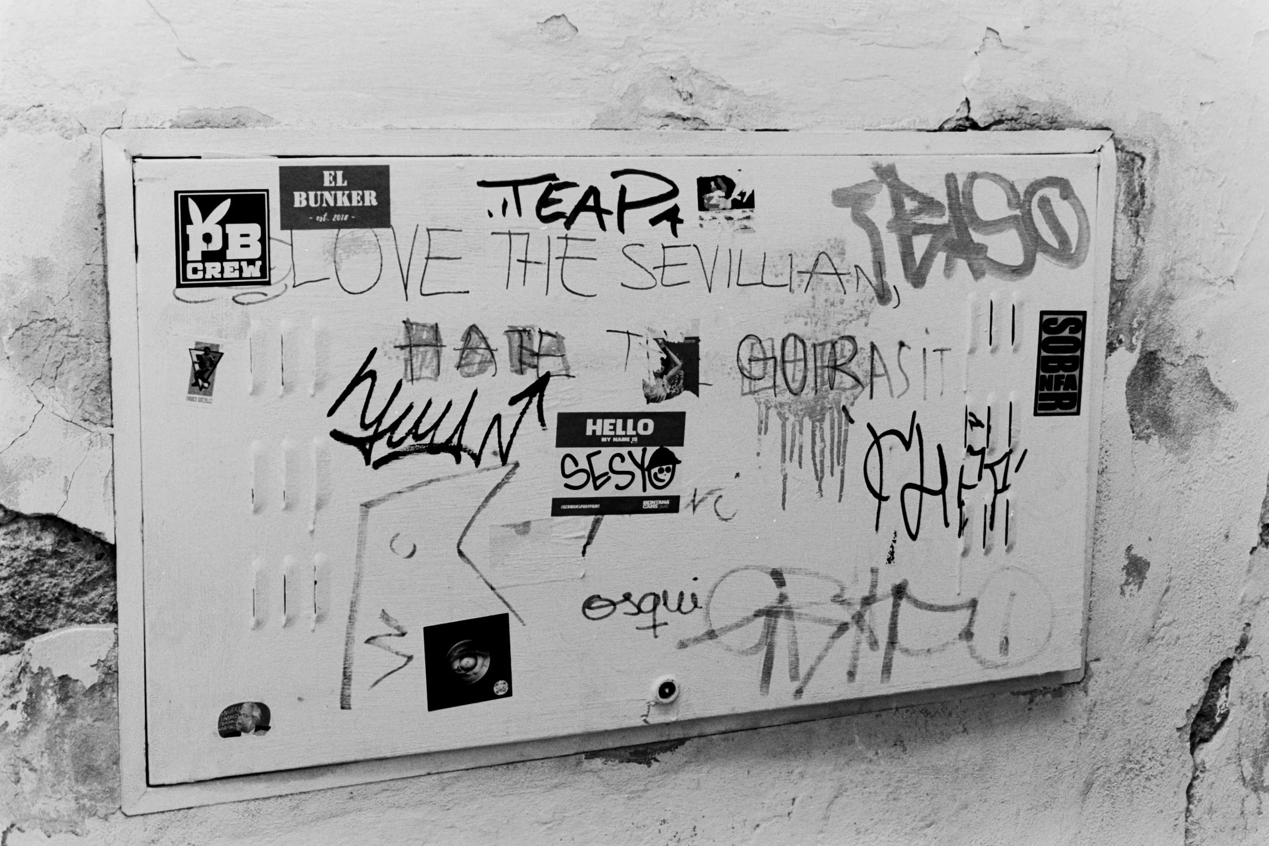 Graffiti-covered bulletin board with stickers and handwritten messages, including 'LOVE THE SEVILLAN', 'HELLO MY NAME IS SESY', and various tags, mounted on a textured stone wall.