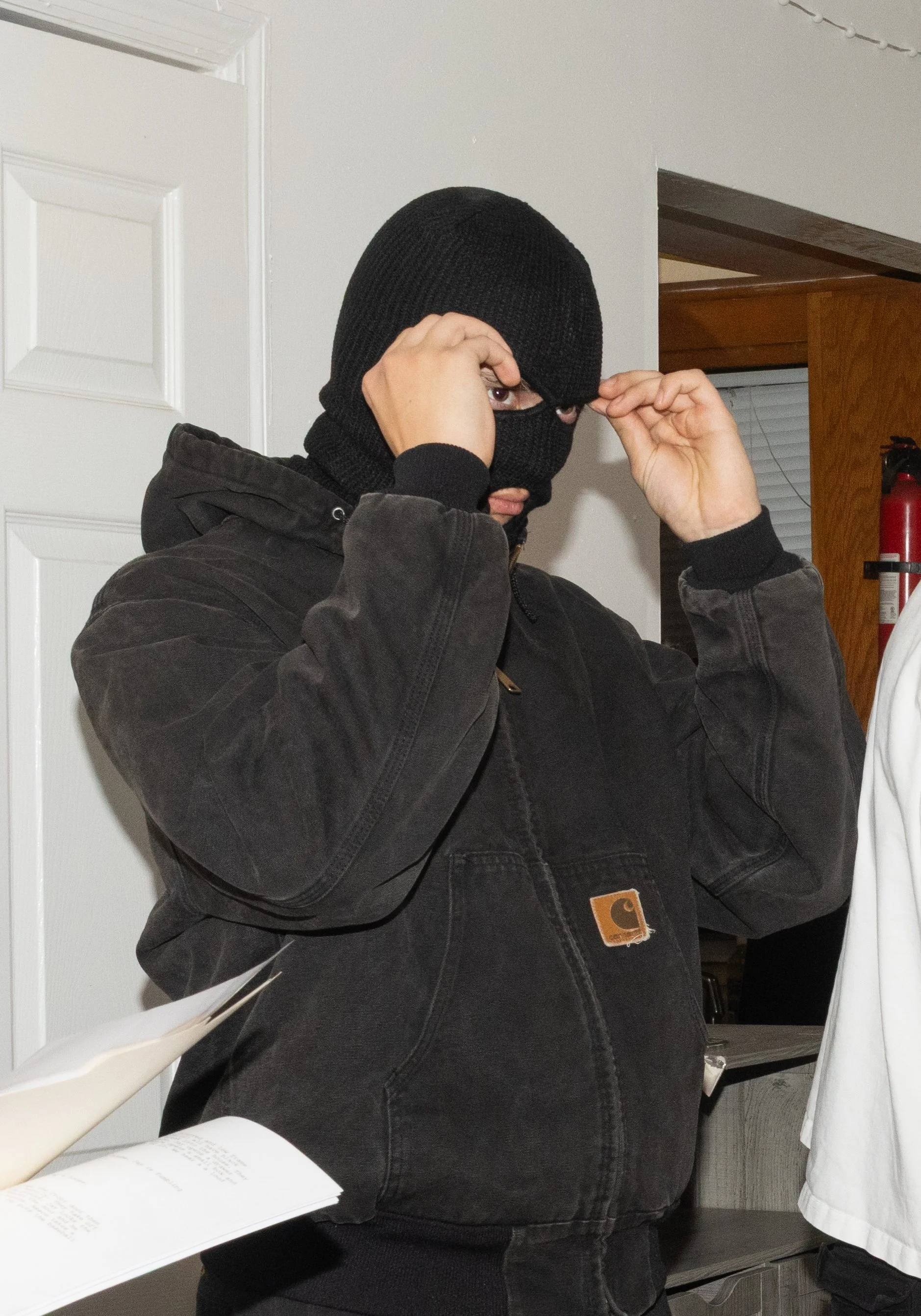 Person wearing a black ski mask and dark Carhartt jacket adjusting the mask in an indoor setting.