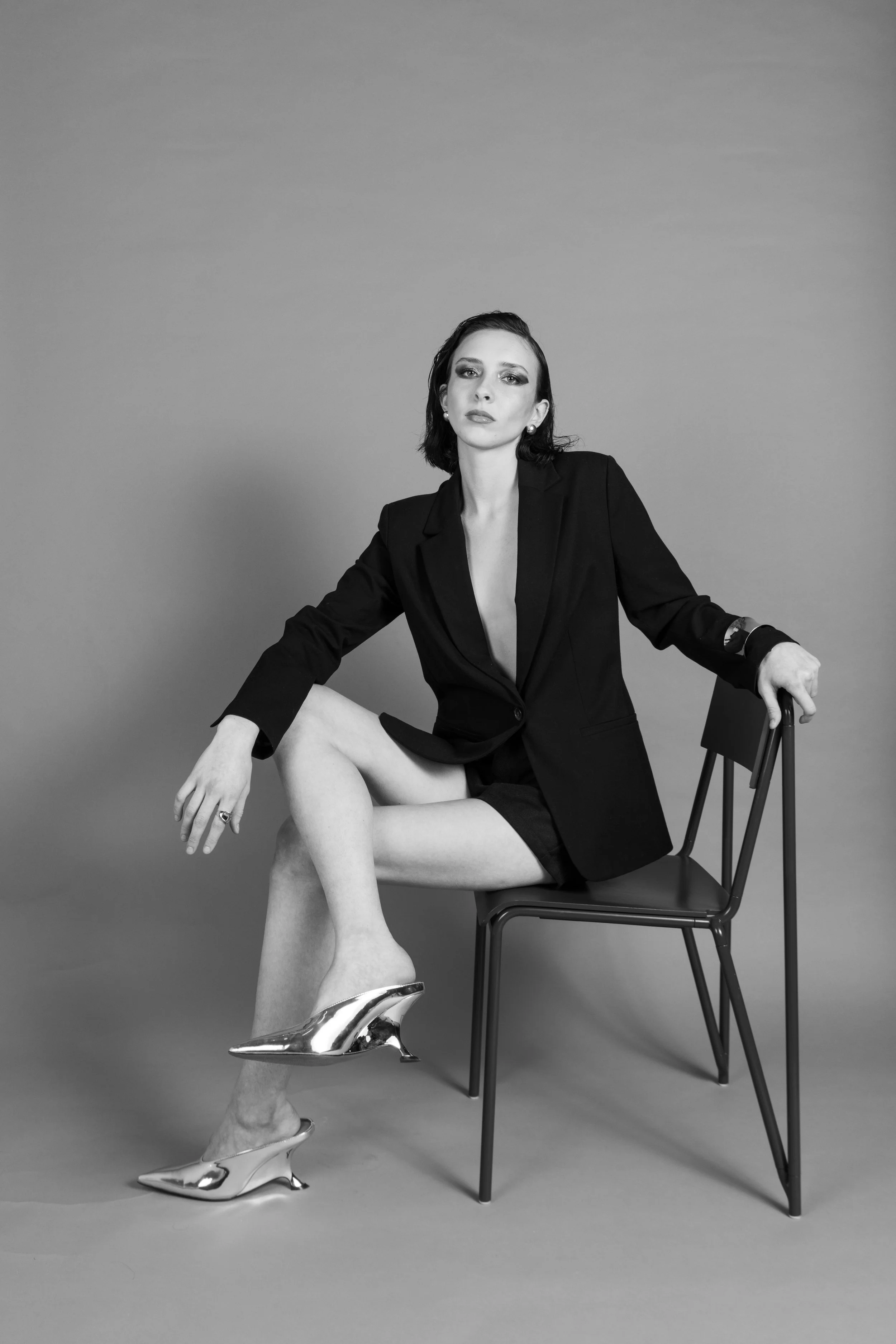 Black-and-white portrait of a woman sitting on a chair, wearing a blazer, shorts, and shiny high heels, with one leg crossed over the other, against a plain background.