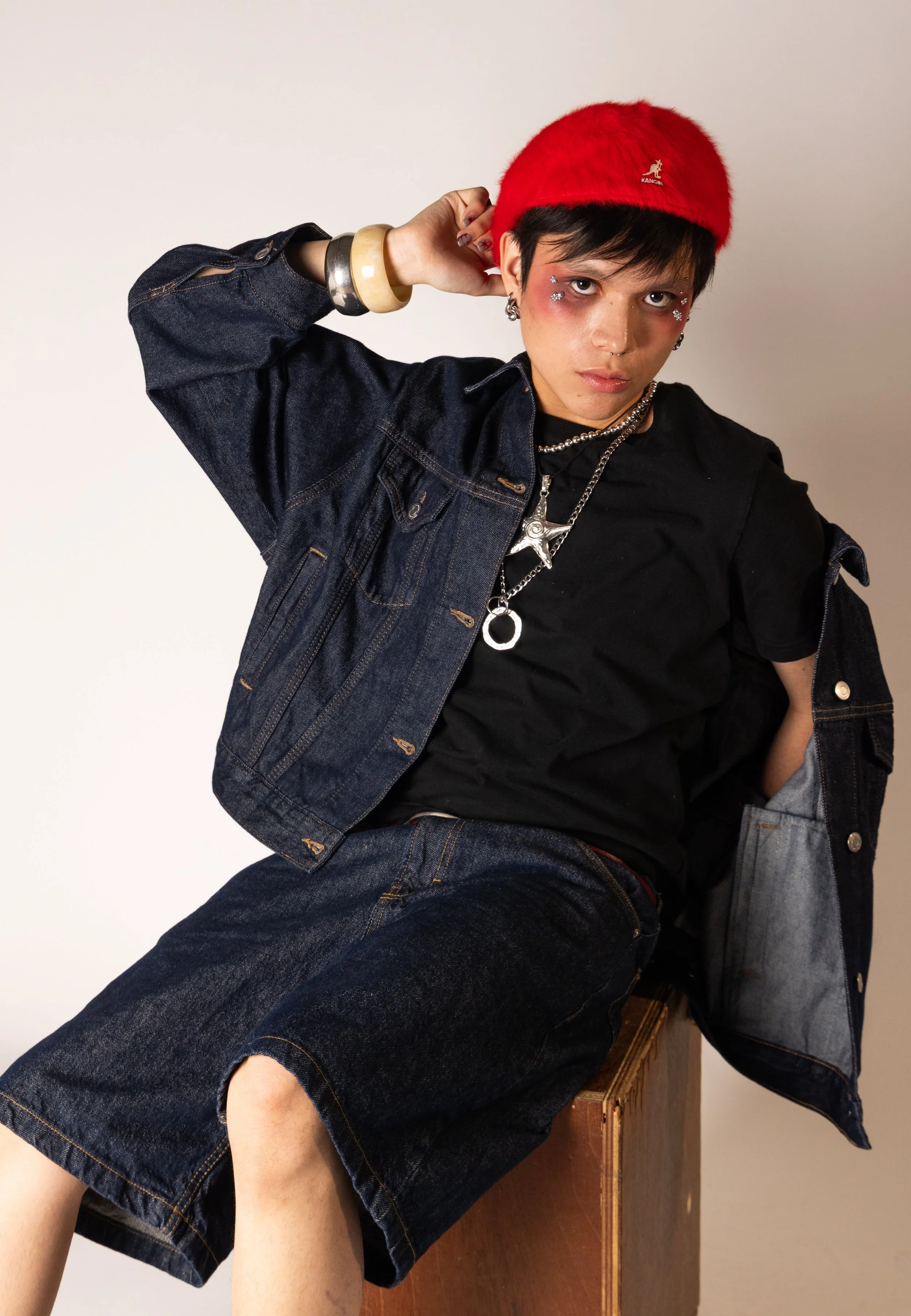 Person with short black hair wearing a red hat, black shirt, denim jacket, and denim skirt, accessorized with necklaces, earrings, and bracelets, sitting on a wooden box against a plain white background.