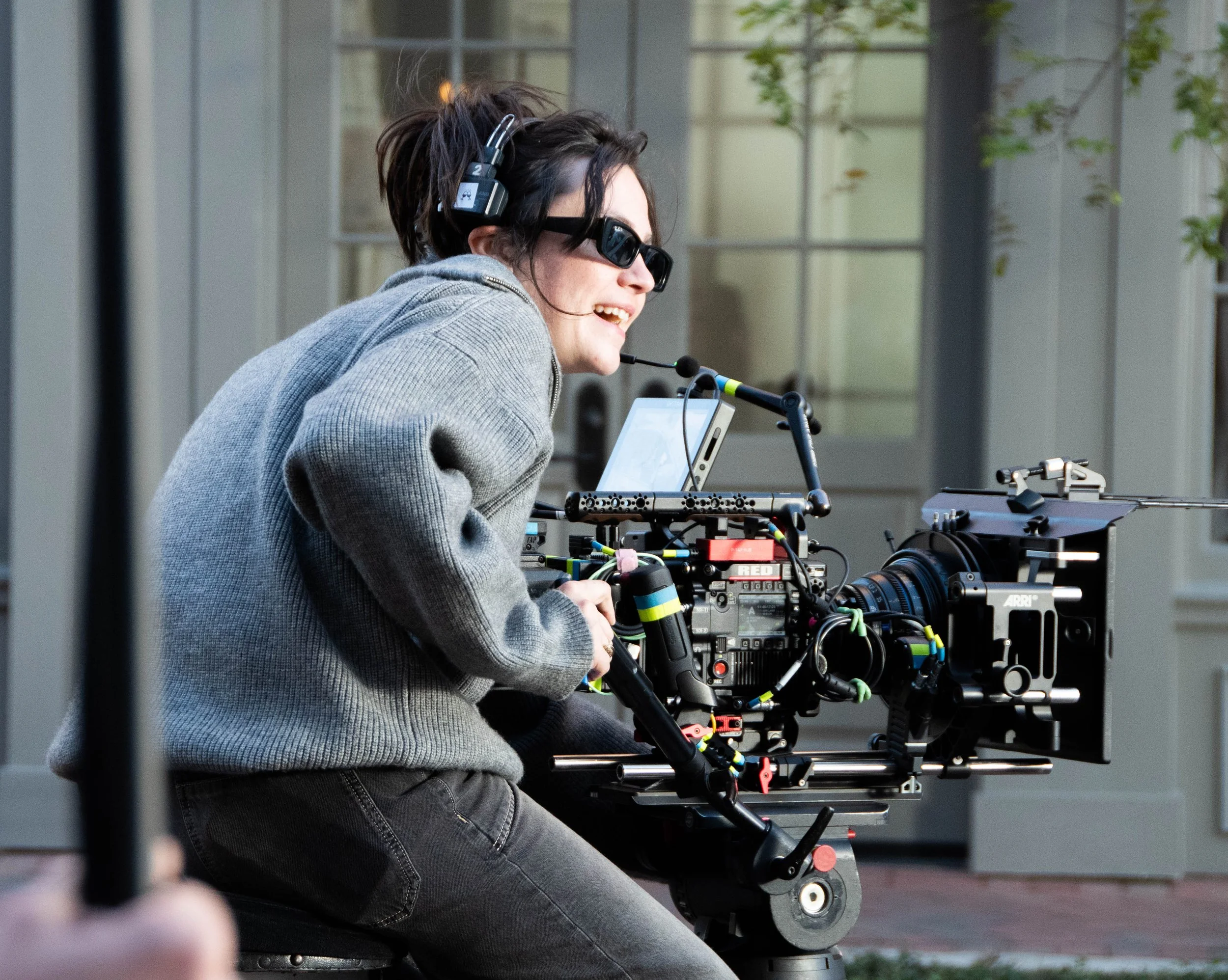 A woman operating a professional film camera outdoors, wearing sunglasses and a headset.