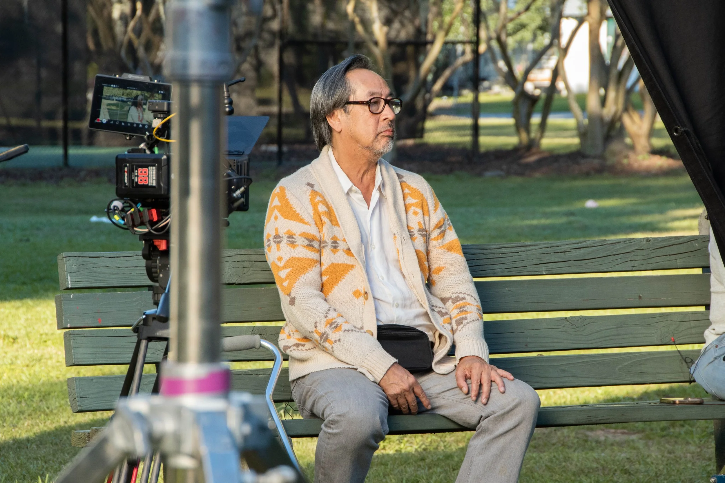 An older man with glasses and gray hair sitting on a park bench during a film shoot. He is wearing a beige cardigan with orange patterns, a white shirt, and gray pants. A camera is set up in front of him, capturing the scene, with filming equipment v