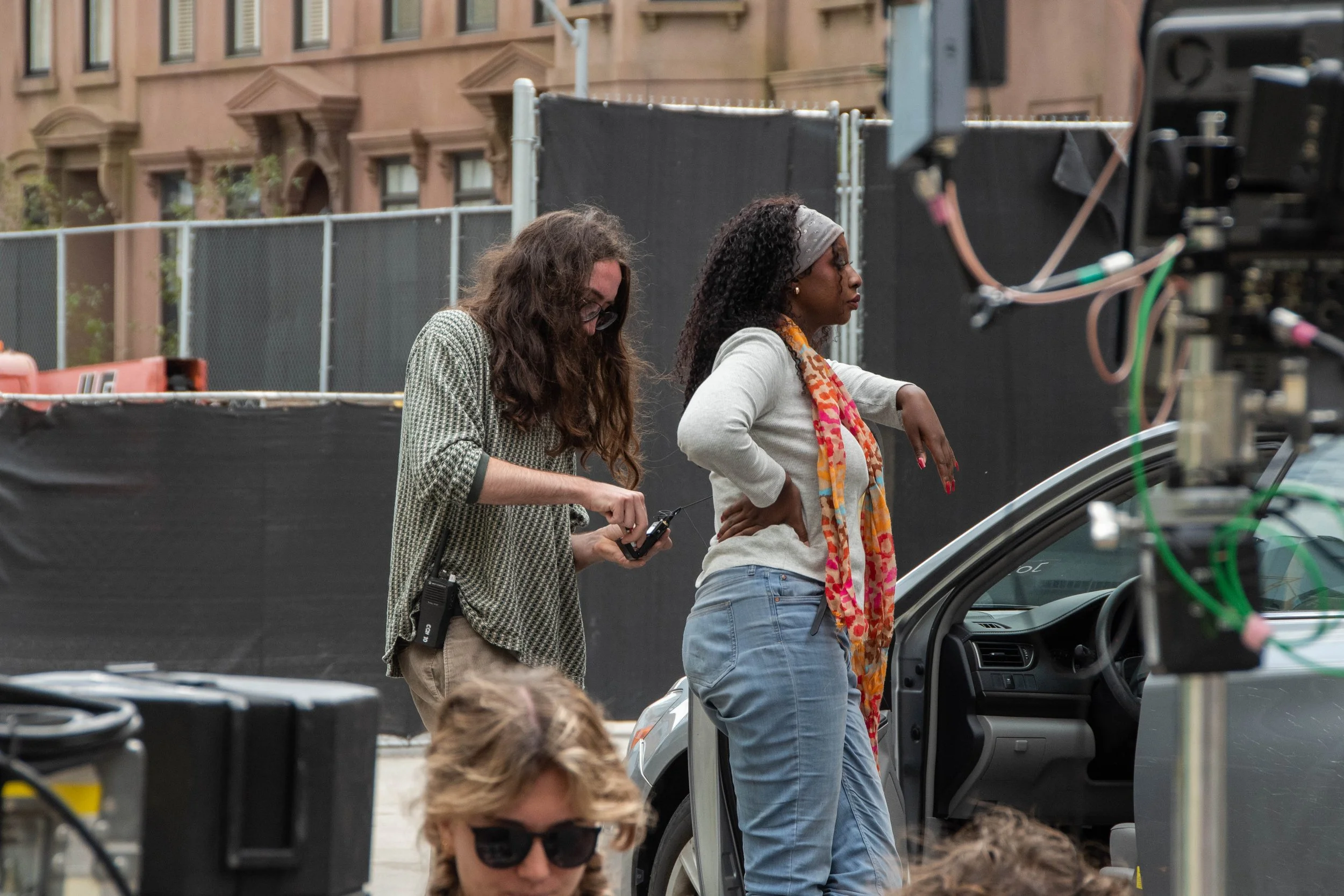 Two women standing next to a car on a film set, one wearing glasses and checking a device, the other with a scarf and hands on hips, with crew and equipment around.