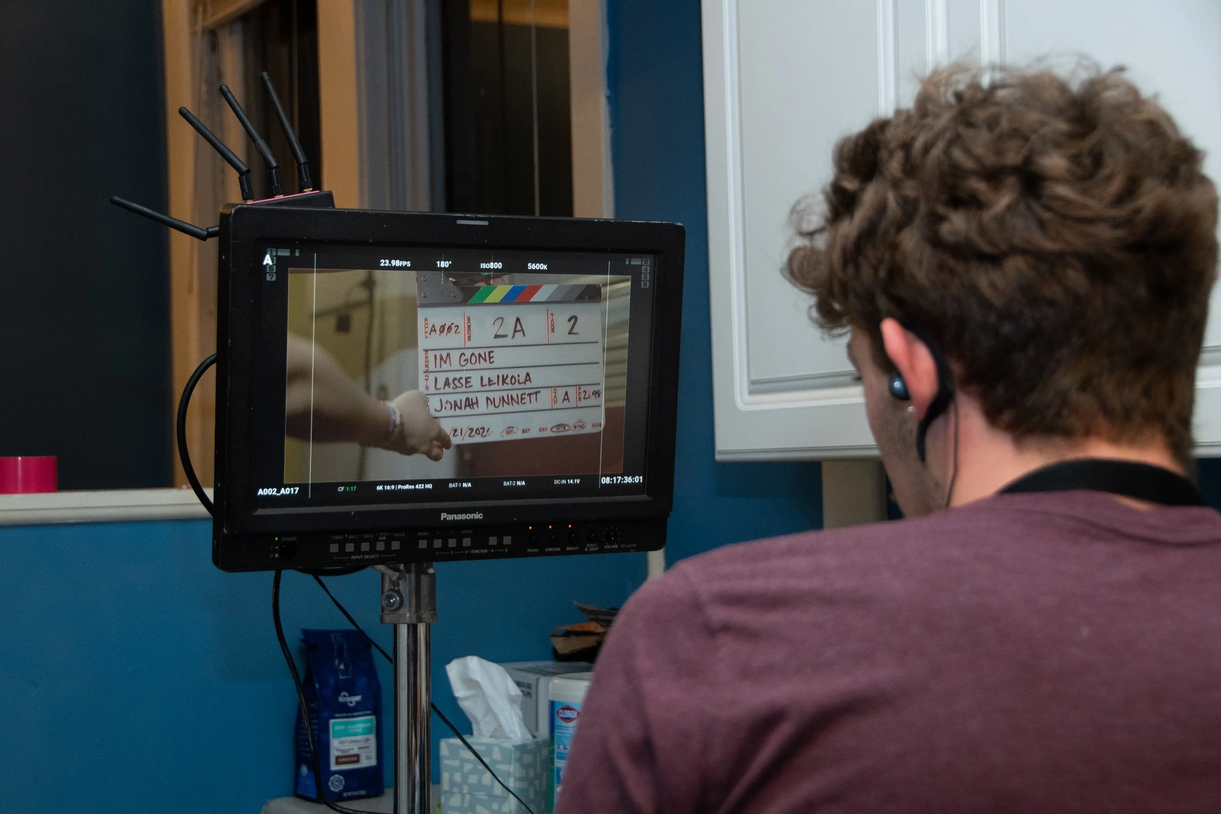 A person with curly hair and wearing a face mask is filming a scene on a monitor, which shows a whiteboard with handwriting and a hand pointing at it. The whiteboard has text including 'IM GONE' and 'JONAH DUNNETT'.