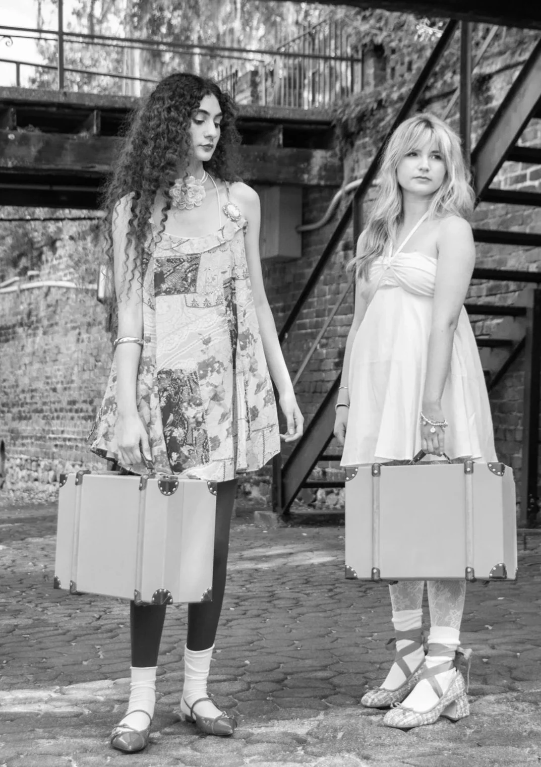 Two young women standing outdoors on a cobblestone street near a brick wall and metal staircase, holding vintage suitcases, dressed in vintage-style clothing, with a serious expression.