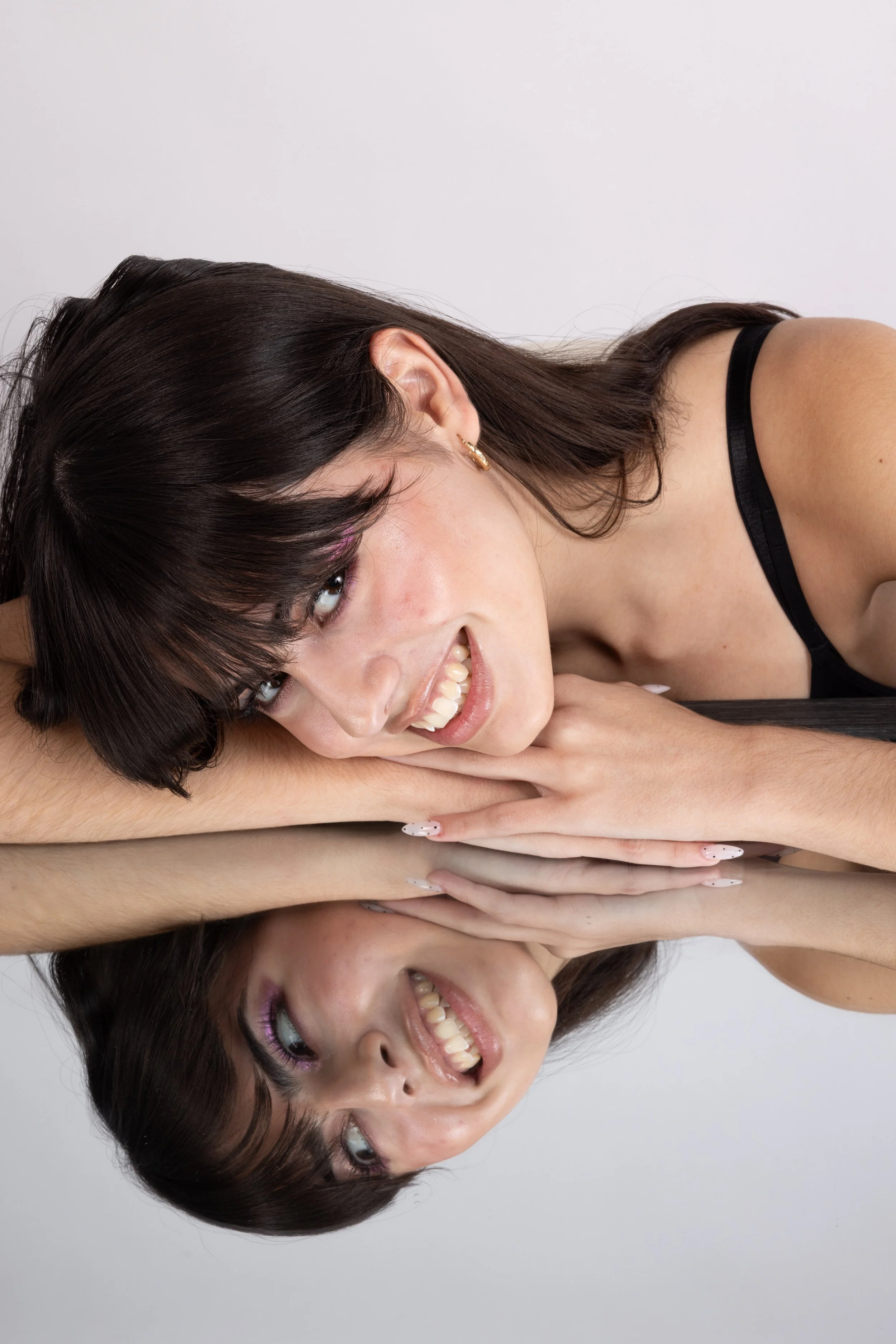 A woman with dark hair and bangs lying on a reflective surface, smiling with her head resting on her hands, creating a mirror image of her face.