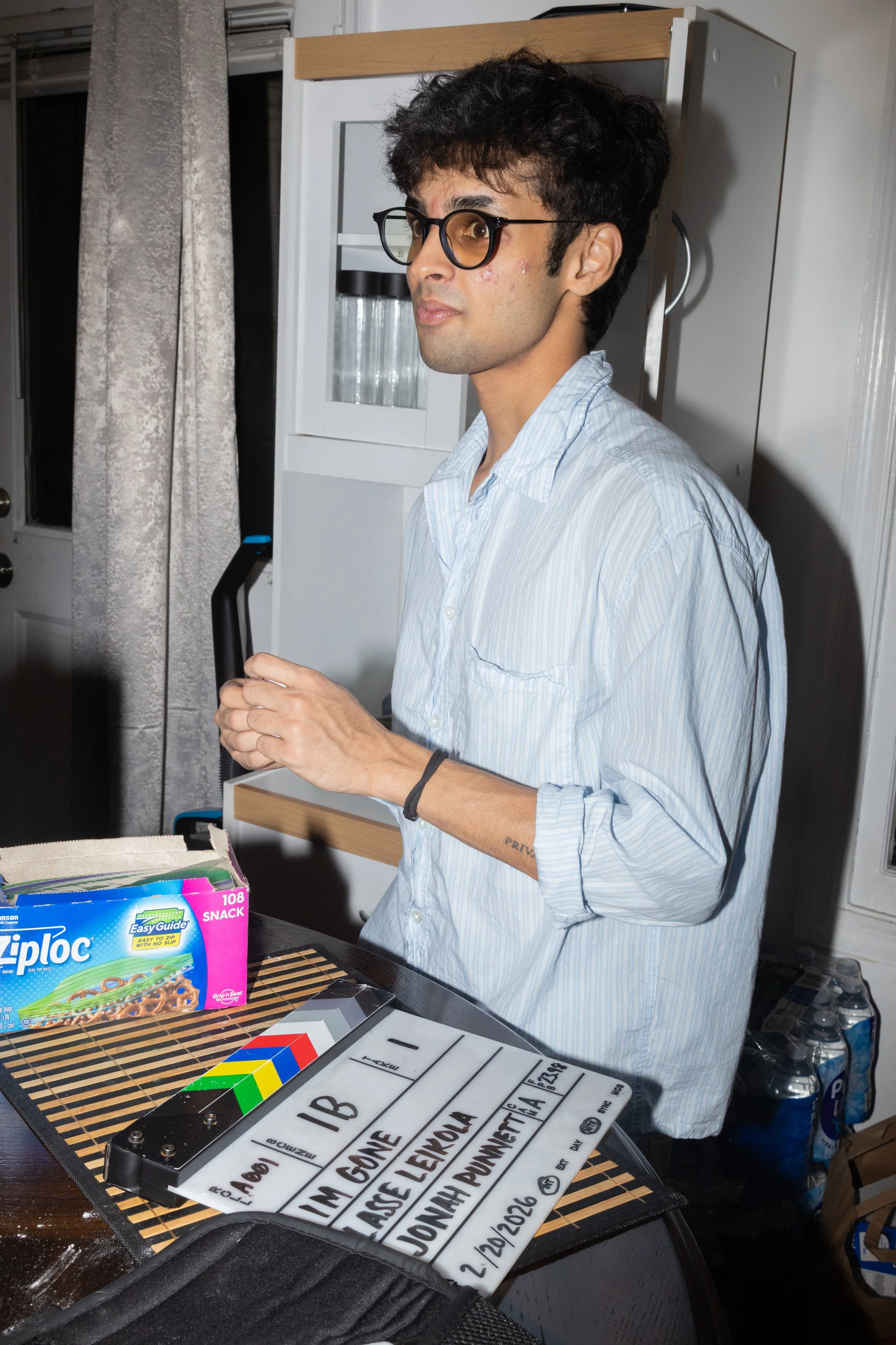 A young man with curly dark hair, glasses, and a light blue button-up shirt standing in a kitchen behind a table, with a Ziploc snack bag, a clapperboard, and some bottles of water visible on the table.