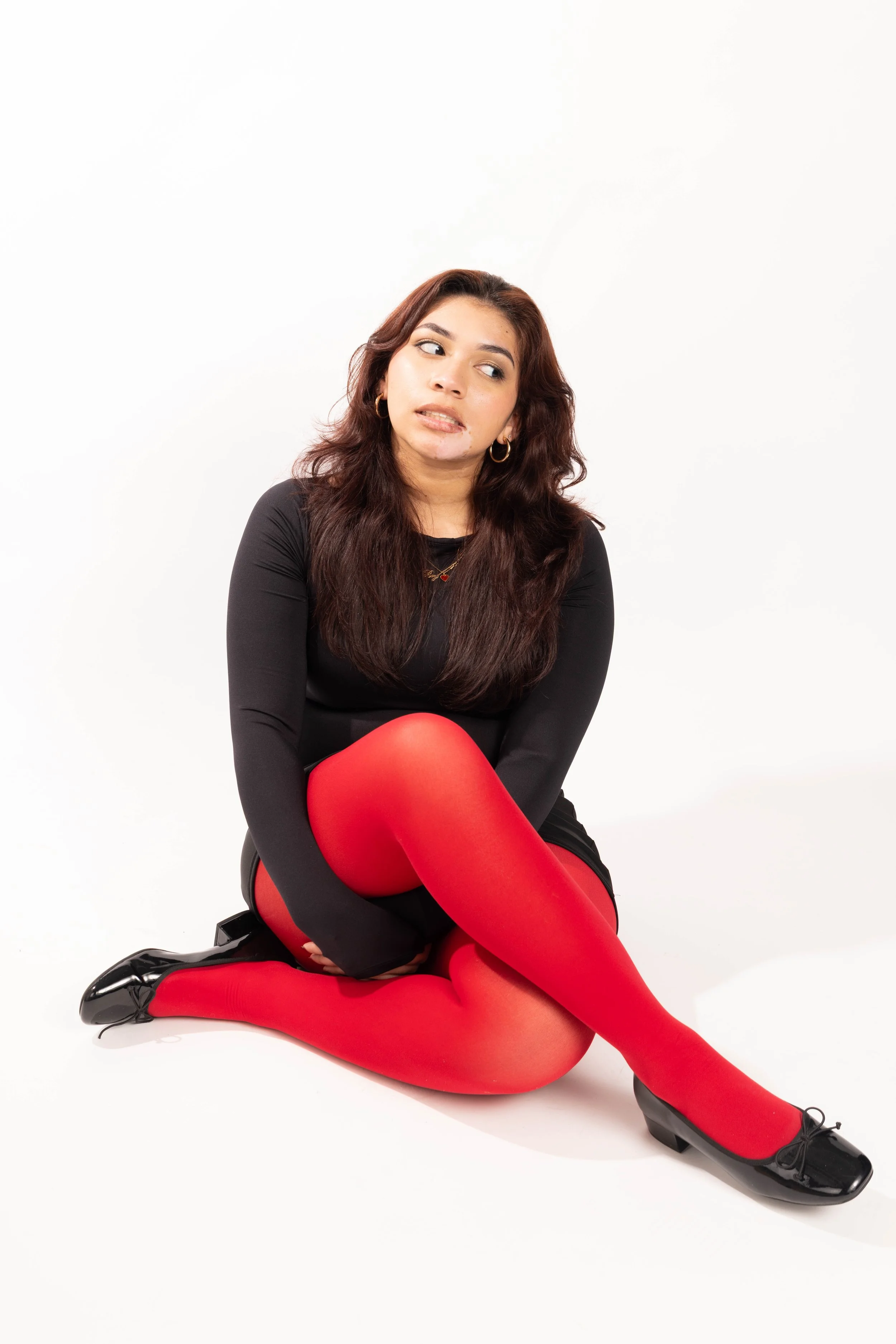 A young woman with long brown hair and hoop earrings sits on the floor in a black dress, red tights, and black shoes, against a plain white background, with an uncertain or contemplative expression.