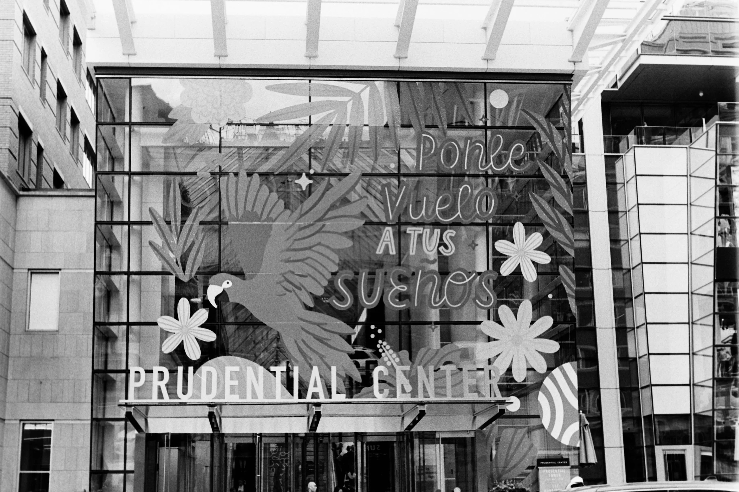 Glass building facade with decorative message and illustrations including a parrot, flowers, and abstract shapes. The message reads "Ponte Velo A TUS SUEÑOS" and "PRUDENTIAL CENTER."