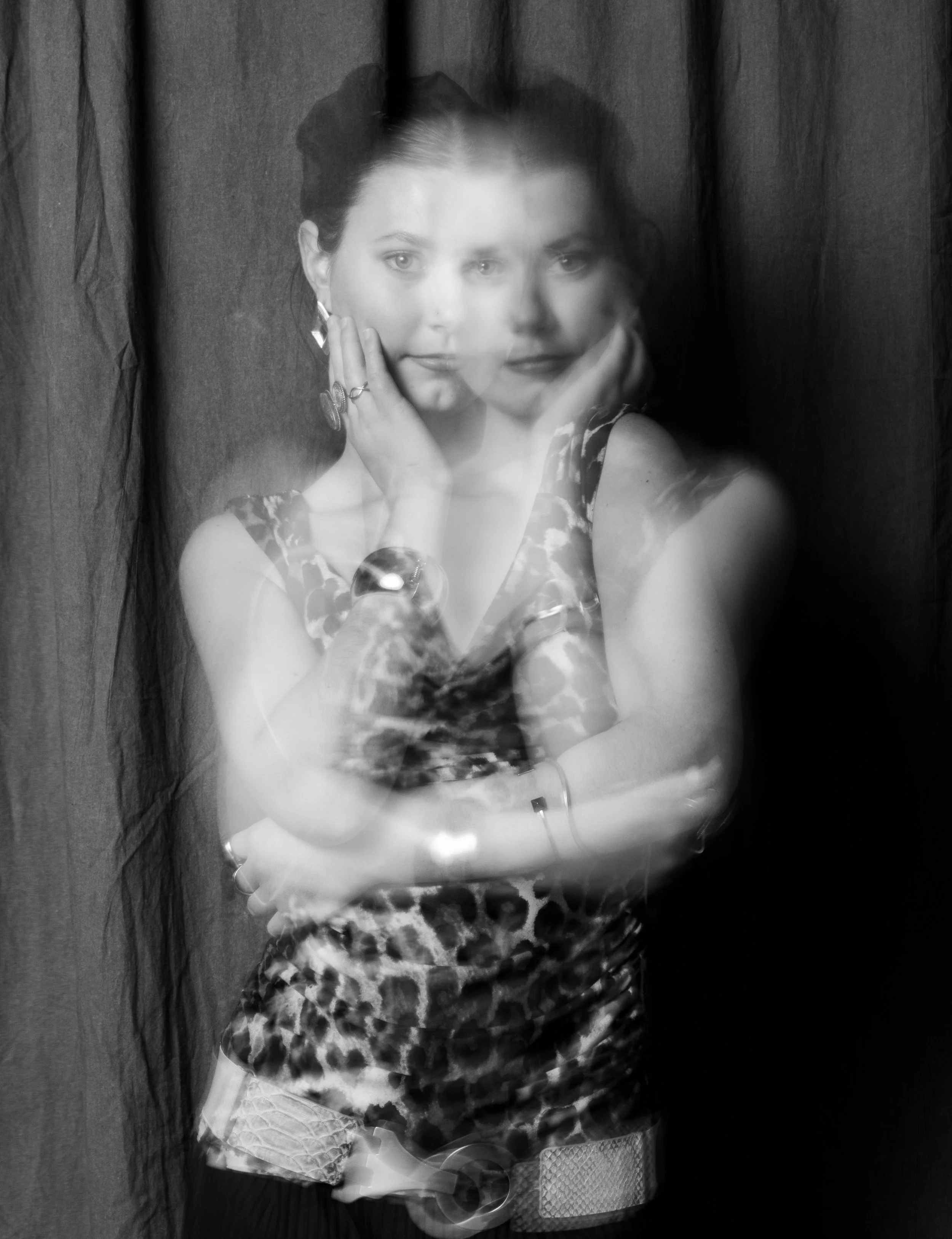 Blurry black and white photo of a woman with long hair, wearing a patterned top and jewelry, posing with her hand on her face against a dark curtain background.