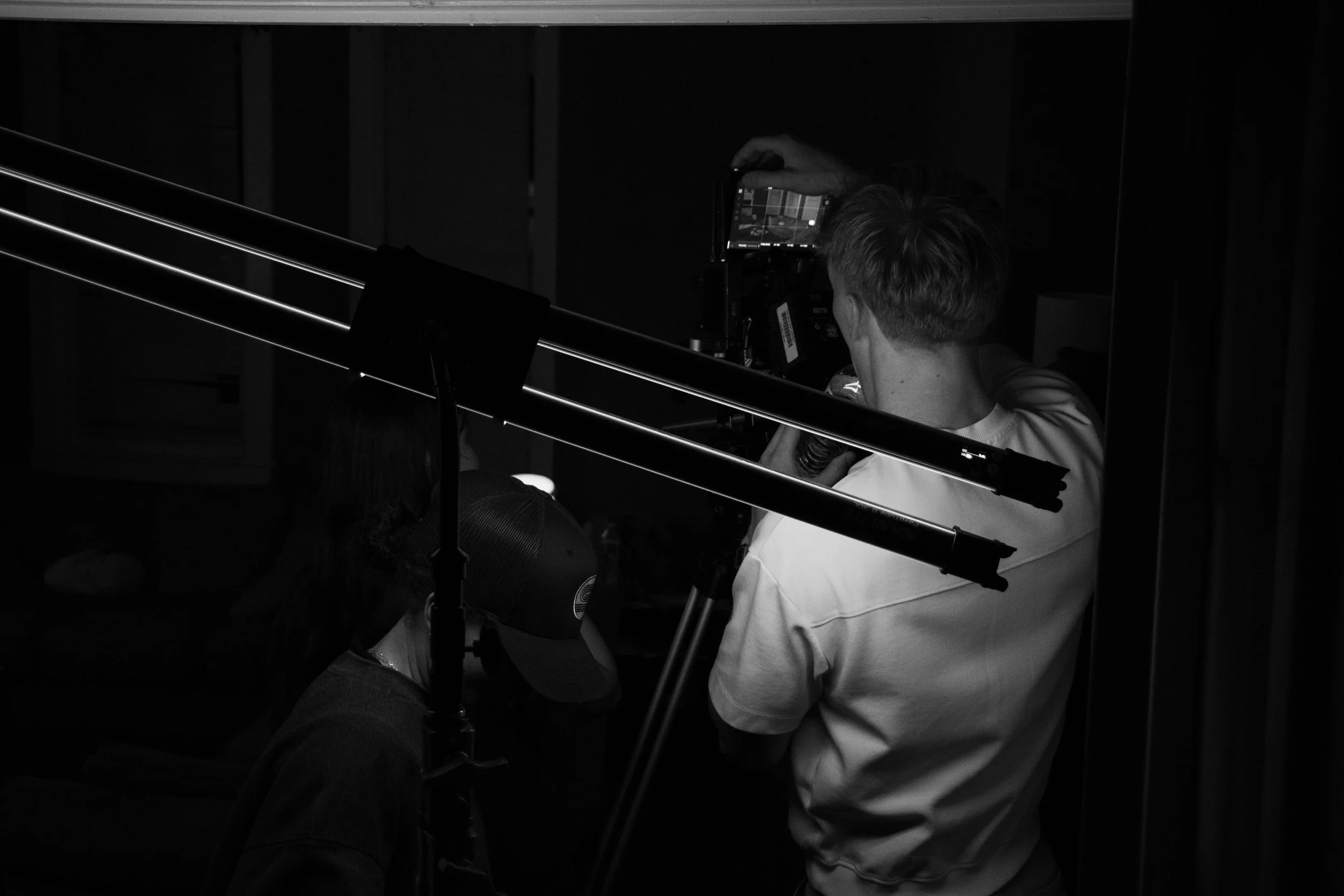 A person operating a camera on a tripod in a dark room, with another person nearby. Overhead lights illuminate the scene.