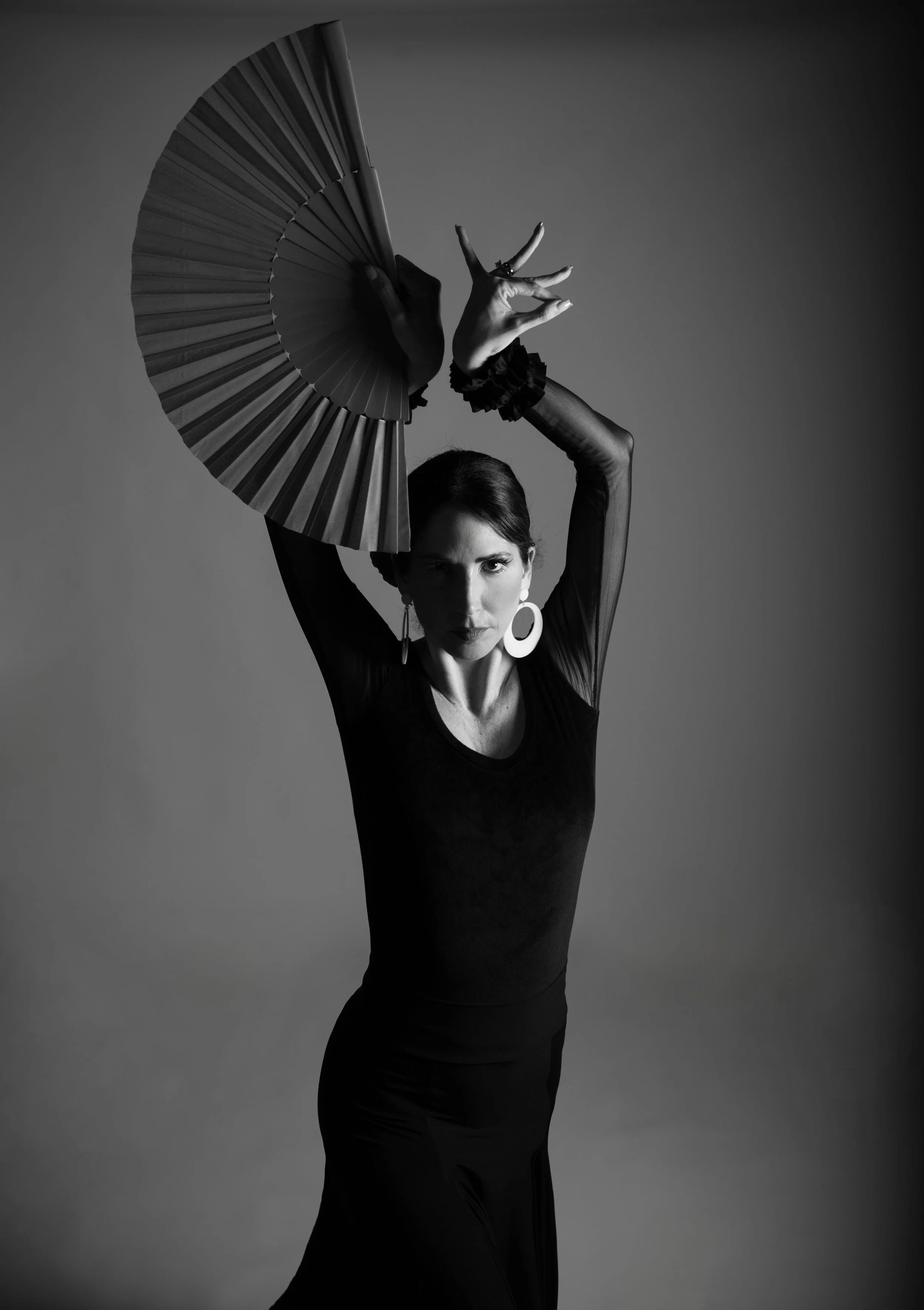 A black and white photo of a woman with dark hair, wearing large earrings and a long black dress, holding a large fan above her head with one hand, striking a dramatic pose.
