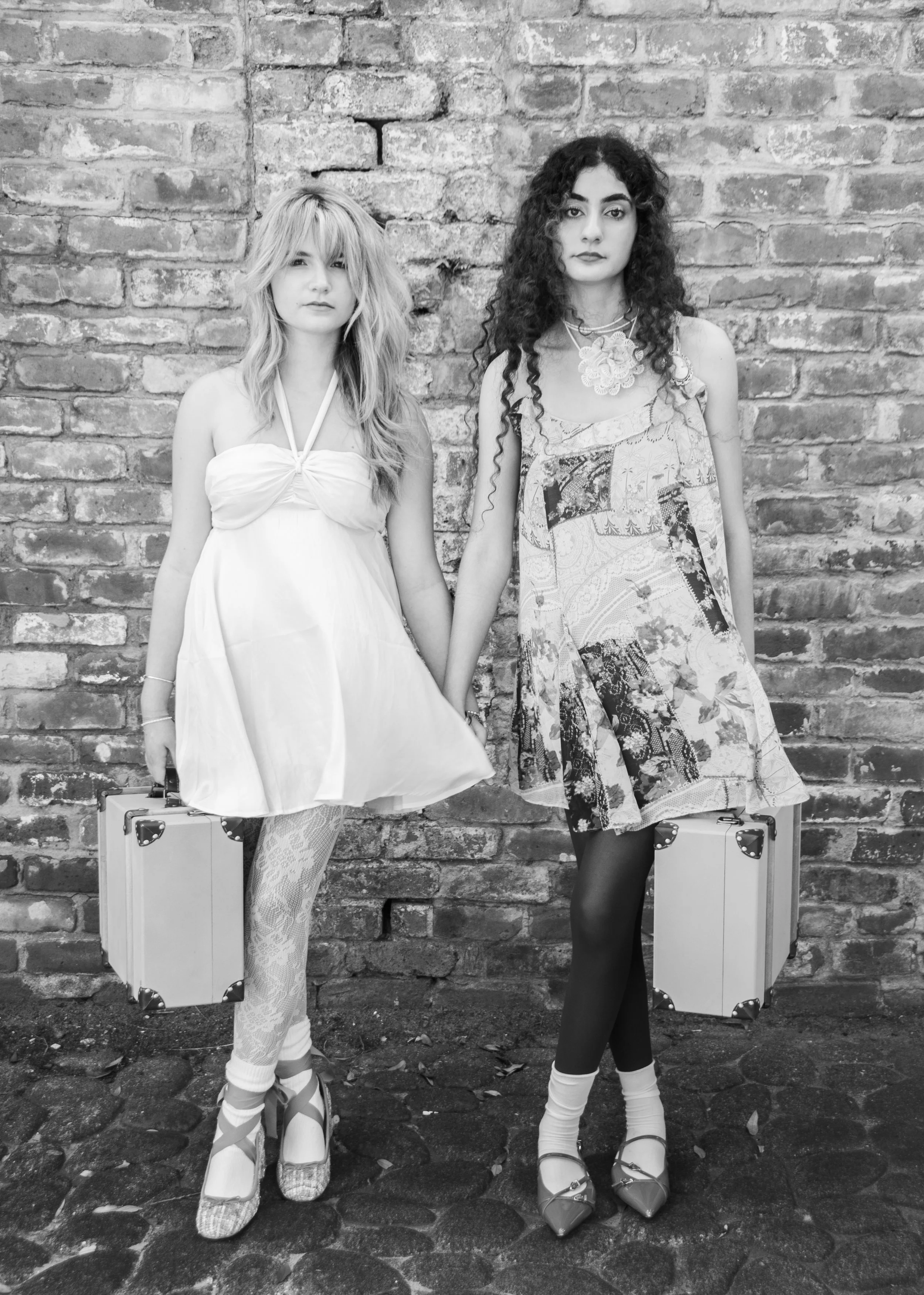 Two young women stand against a brick wall, holding hands and carrying vintage-style suitcases. One wears a short, strapless dress with lace tights and high heels, while the other wears a patterned dress with tights and heels. Both have curly hair an