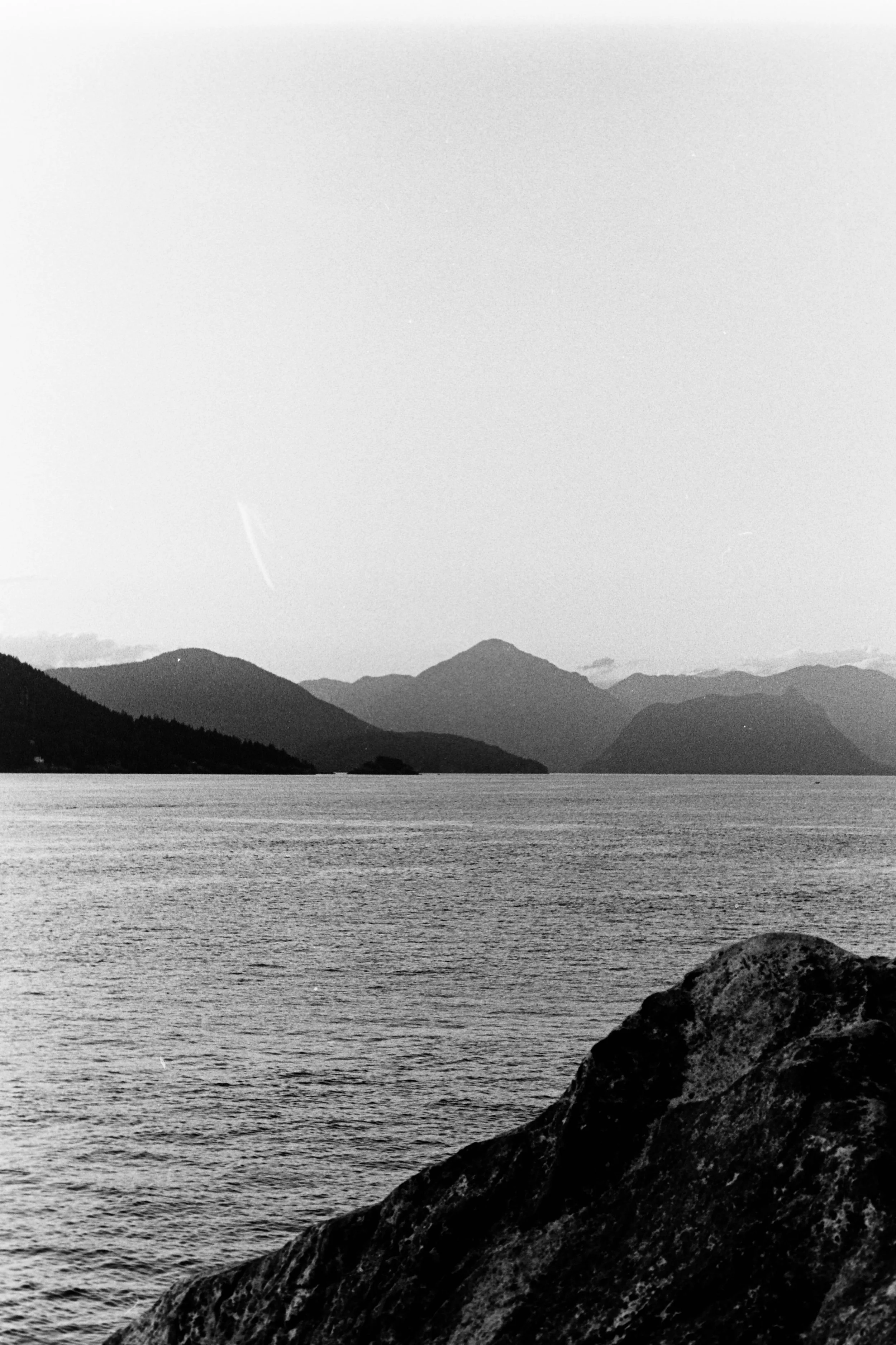 Black and white image of a large body of water with mountains in the background and a large rock in the foreground.