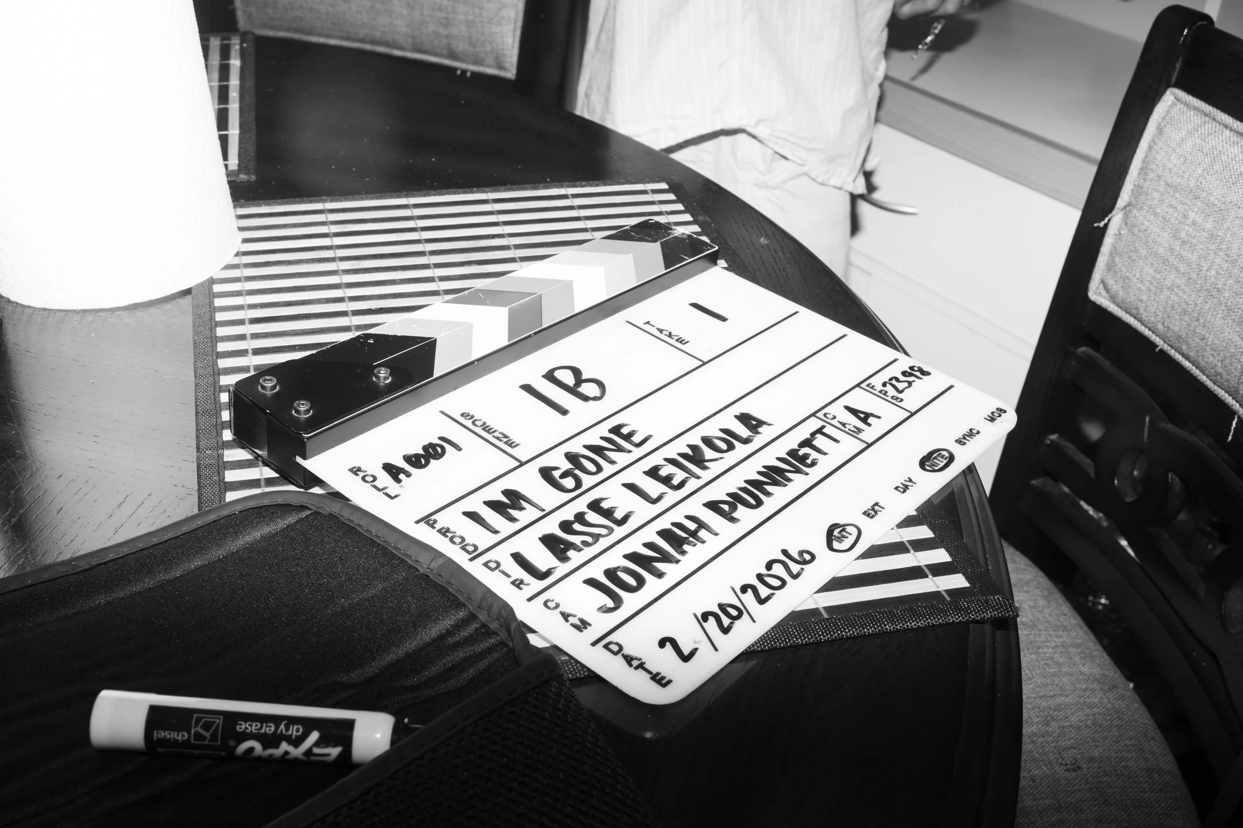 A film slate with handwritten details placed on a dining table, next to a black bag containing a white marker, with chairs and a window visible in the background.