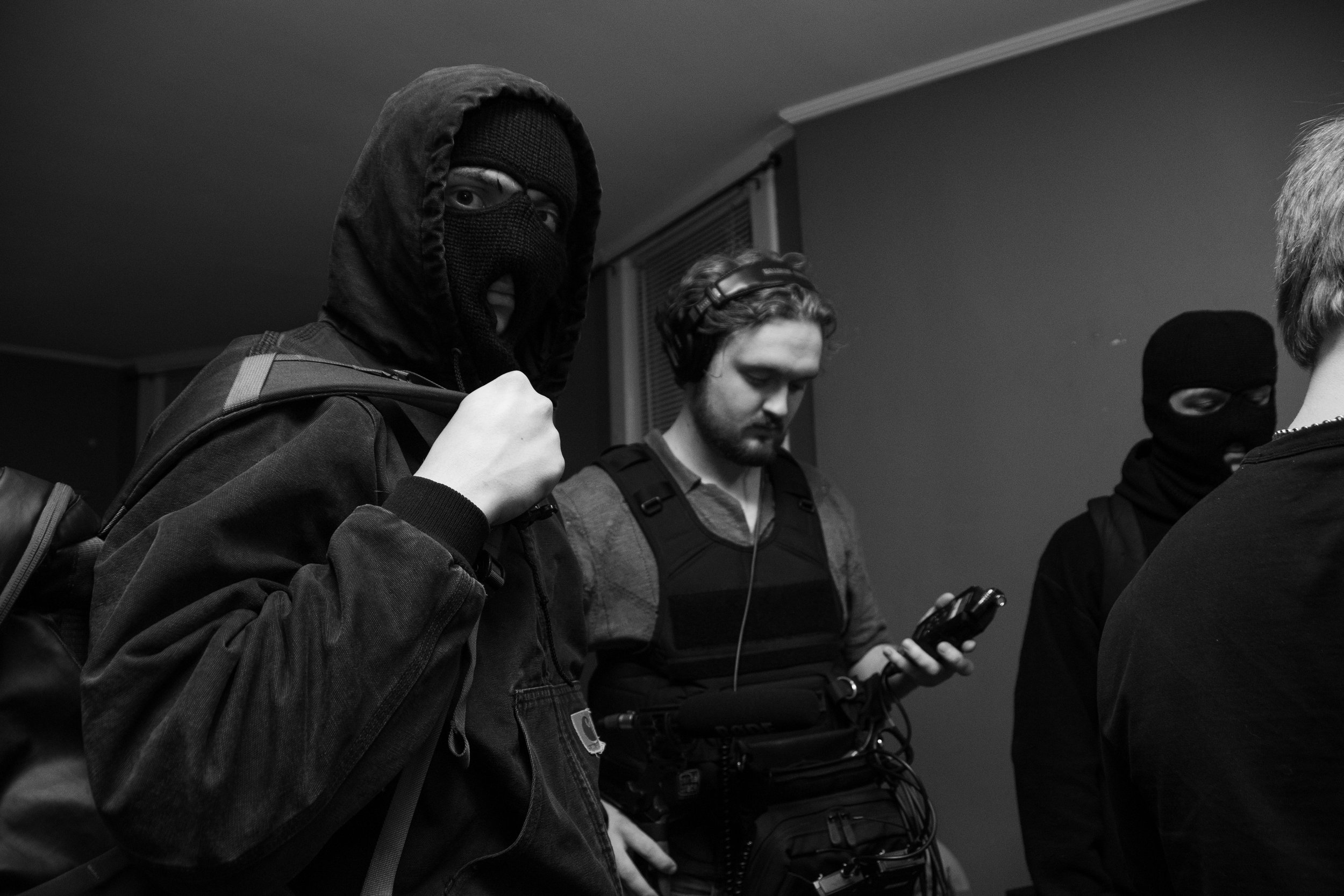 Black and white photo of two masked men in hooded sweatshirts and a man with headphones and a camera gear, standing indoors.