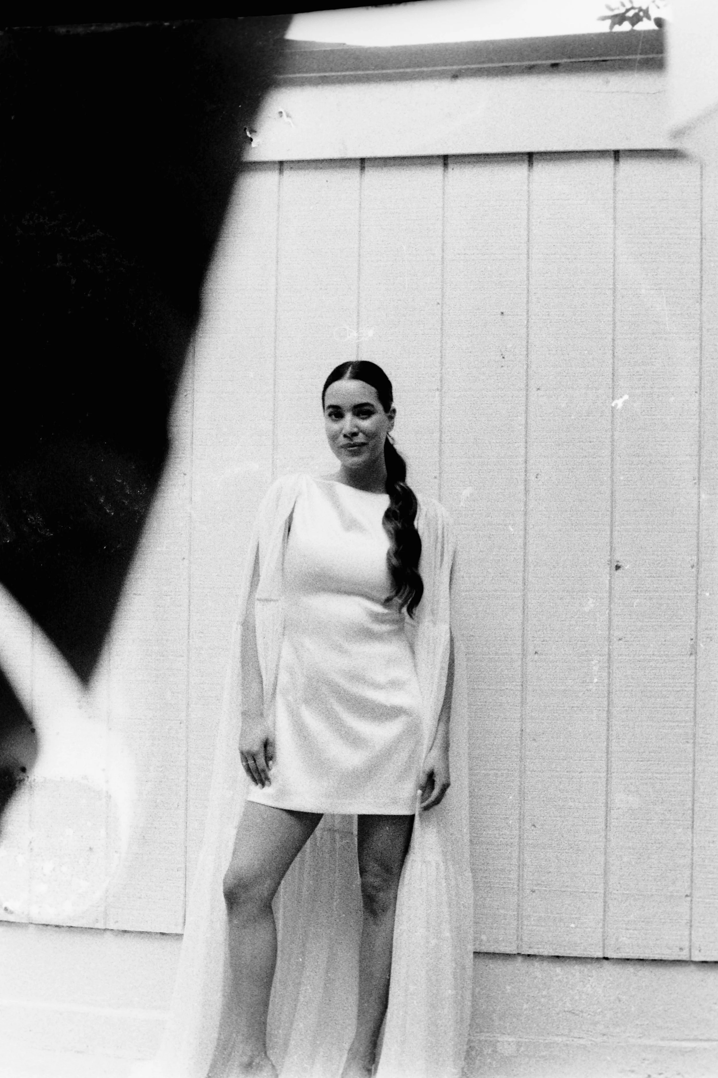 Black and white photo of a woman with long hair styled in a braid, standing outdoors in front of a wooden wall, wearing a light-colored dress.