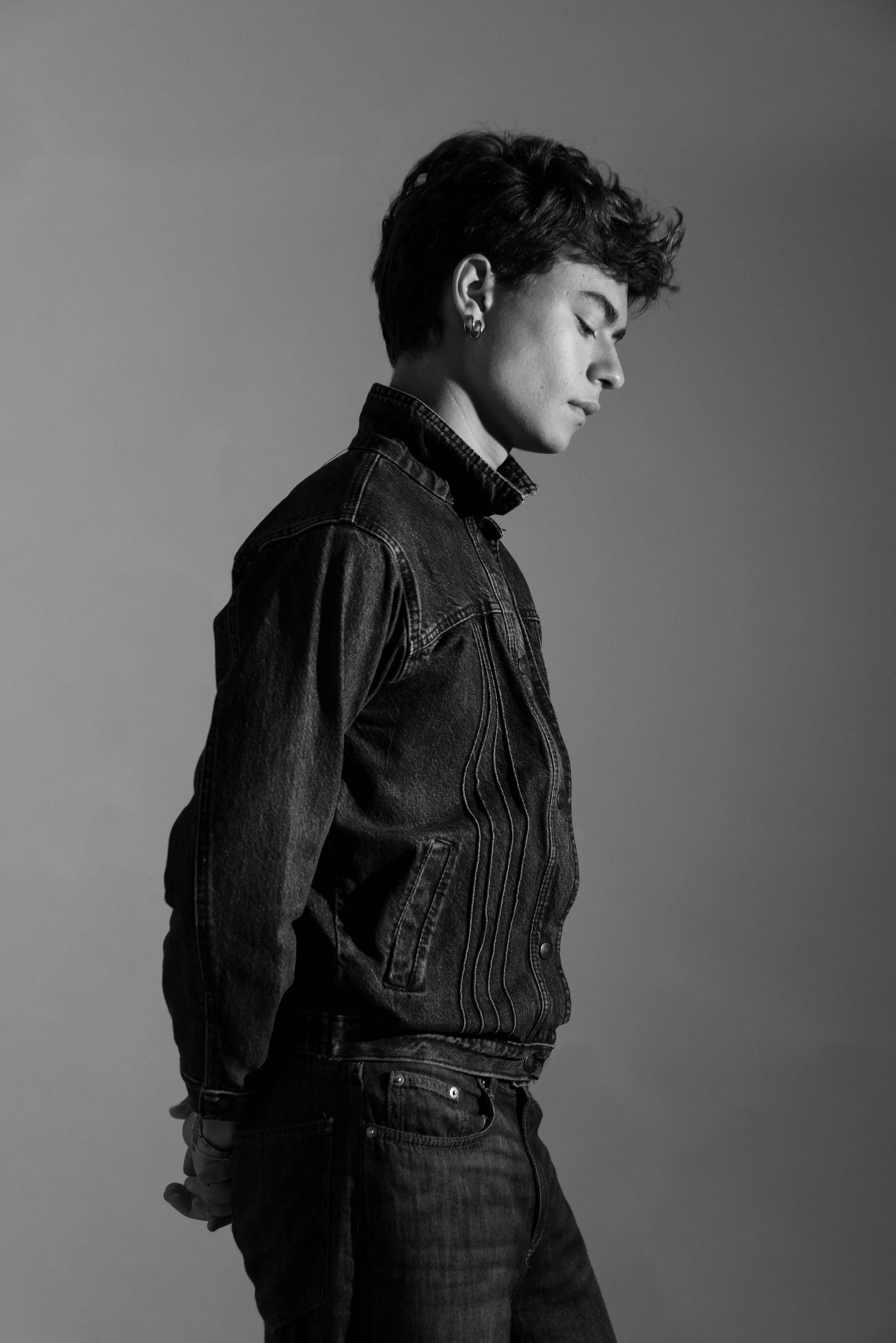 A black and white photo of a young person with short, wavy hair, wearing a denim jacket and pants, standing in profile against a plain background.