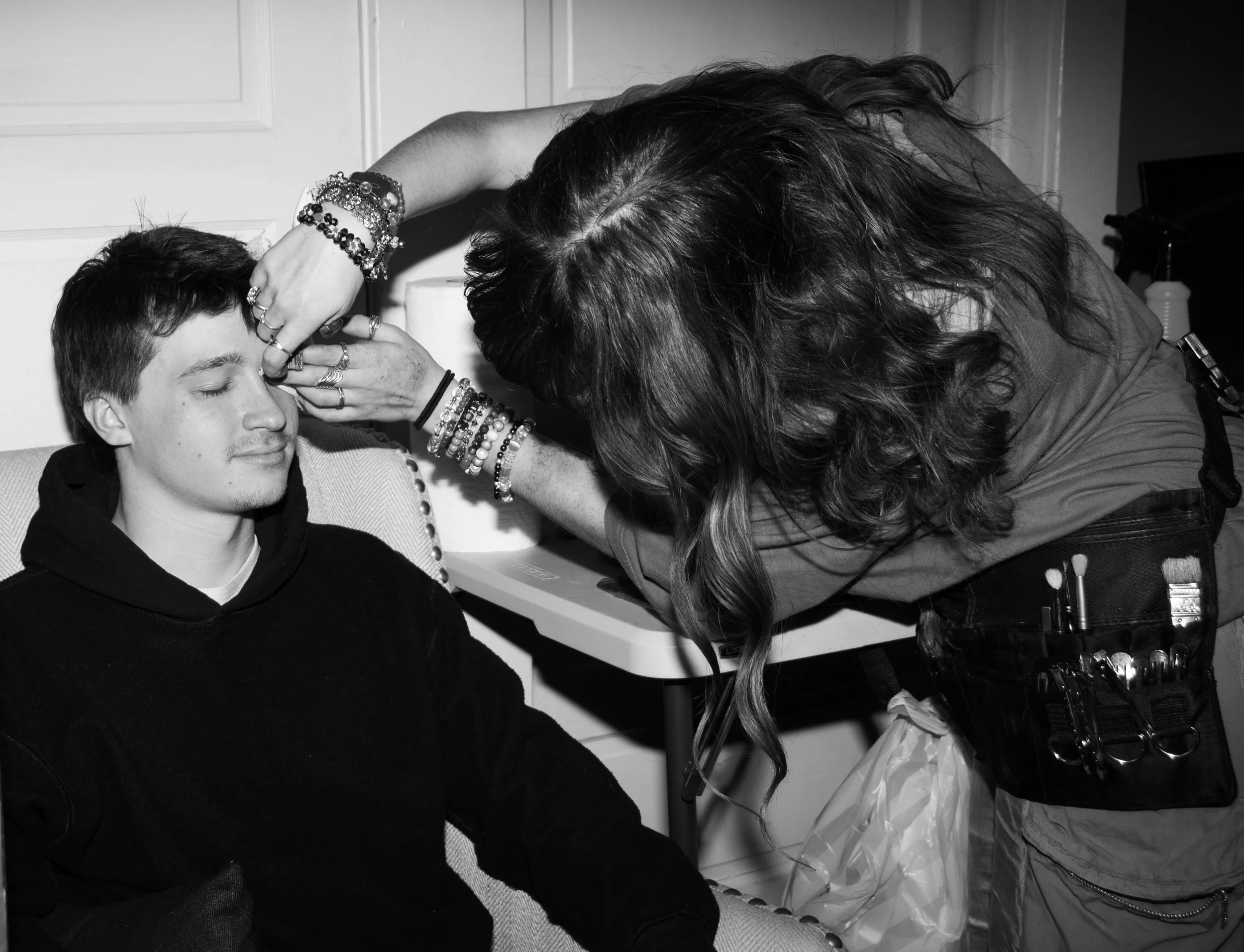 A woman applying makeup to a young man with long hair, who is seated with eyes closed, in a room with a white door and a table.