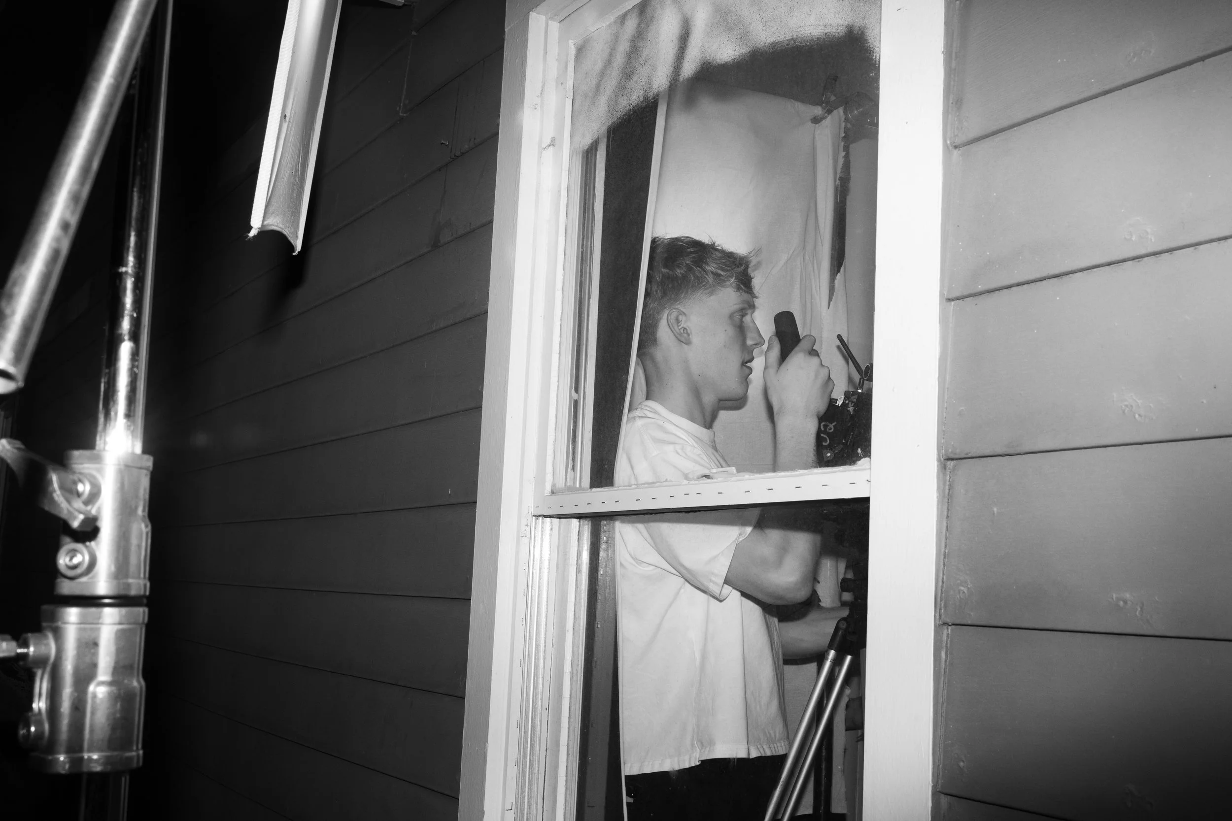 A young man using a camera through a window on the side of a house.