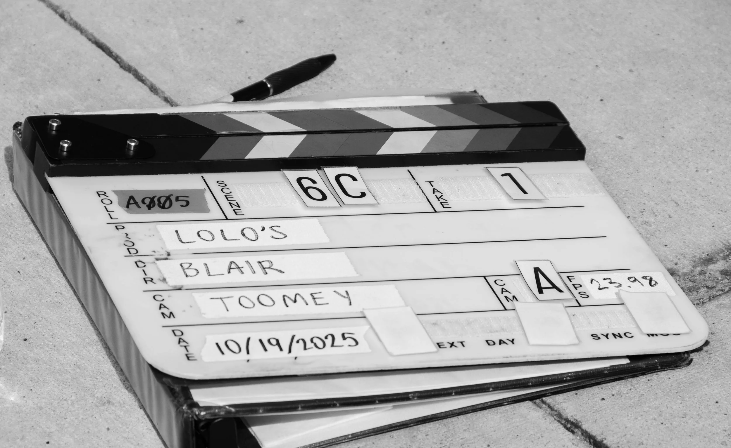 A clapperboard on a surface with a pen resting on it. The clapperboard has information written on it, including names, date, and scene details.