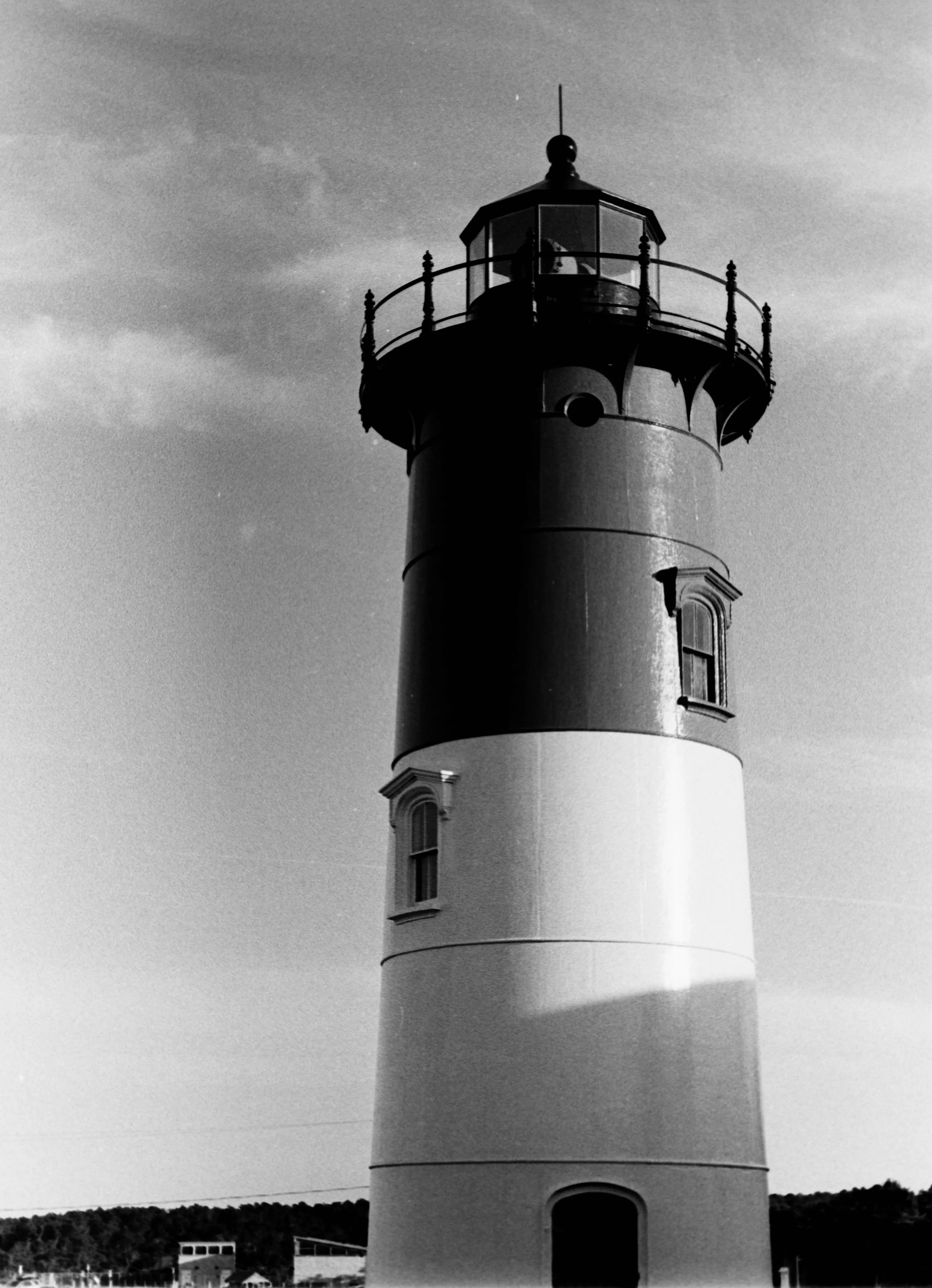 A black and white photo of a tall lighthouse with a round top and balcony, against a cloudy sky.