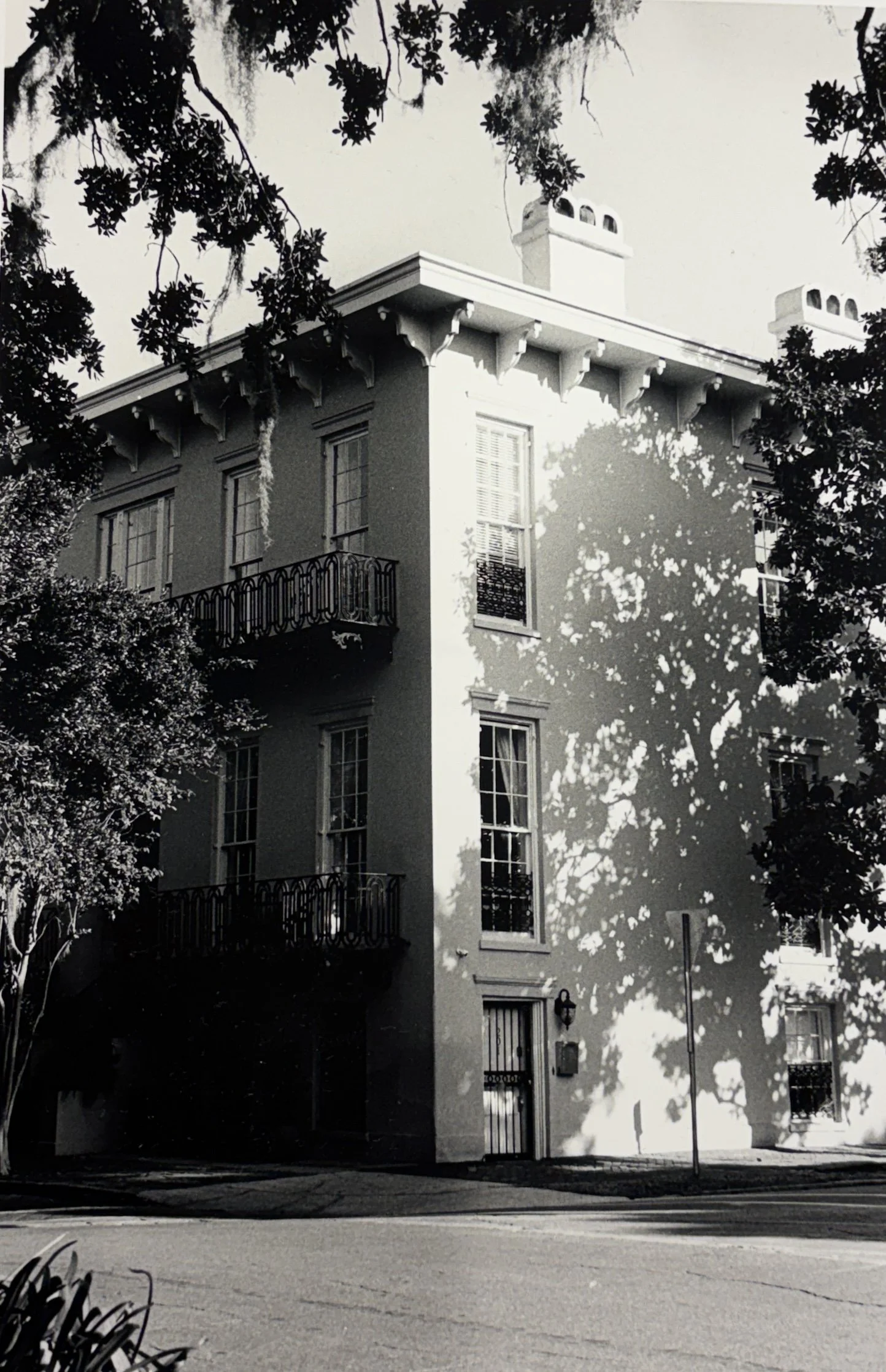 Black and white photo of a corner multi-story building with balconies, trees casting shadows on the facade, and a street scene with a street sign and lamp post.