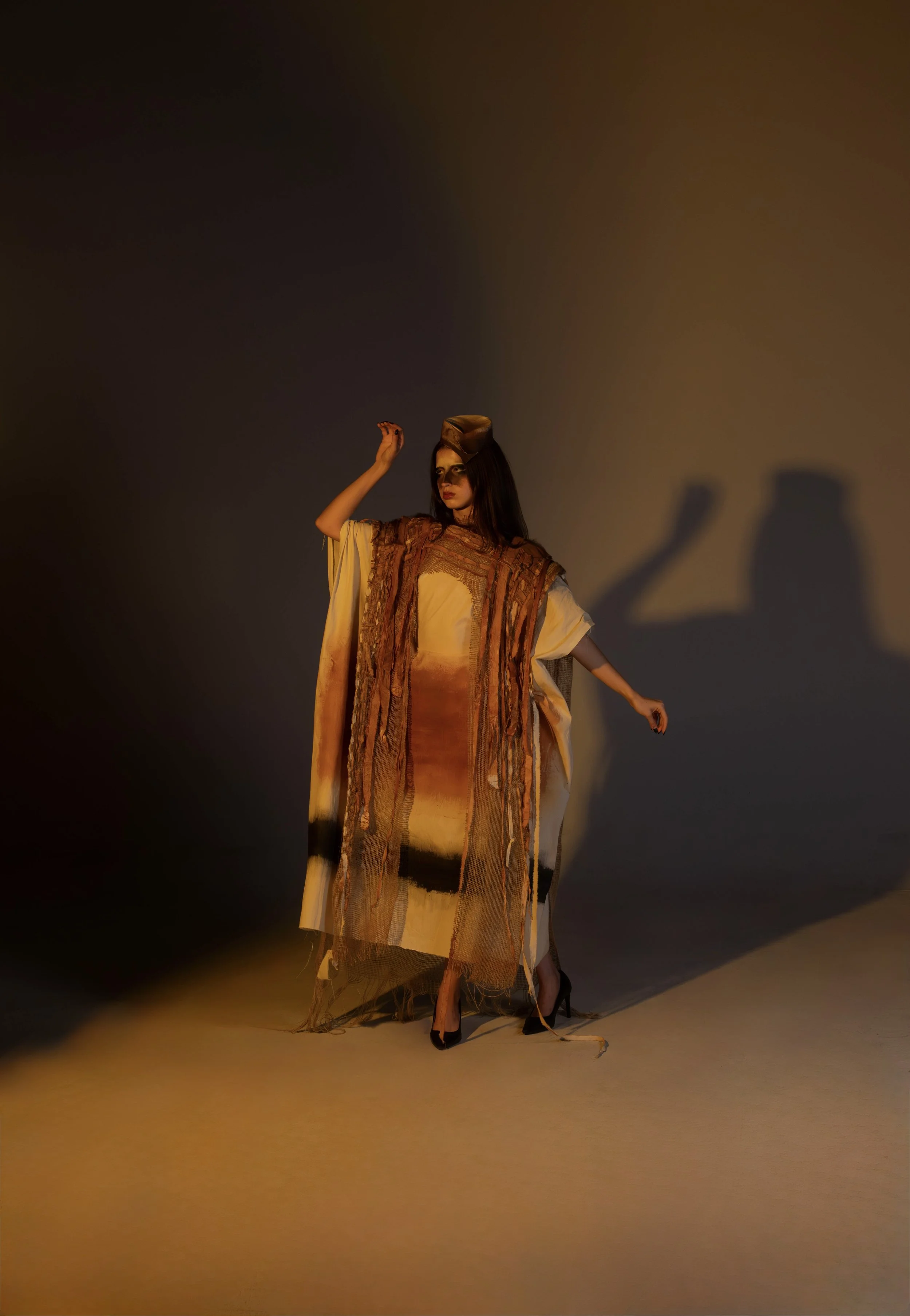 A woman wearing a multicoloredly painted dress and a brown headscarf, standing against a dark background with shadows cast on the wall.