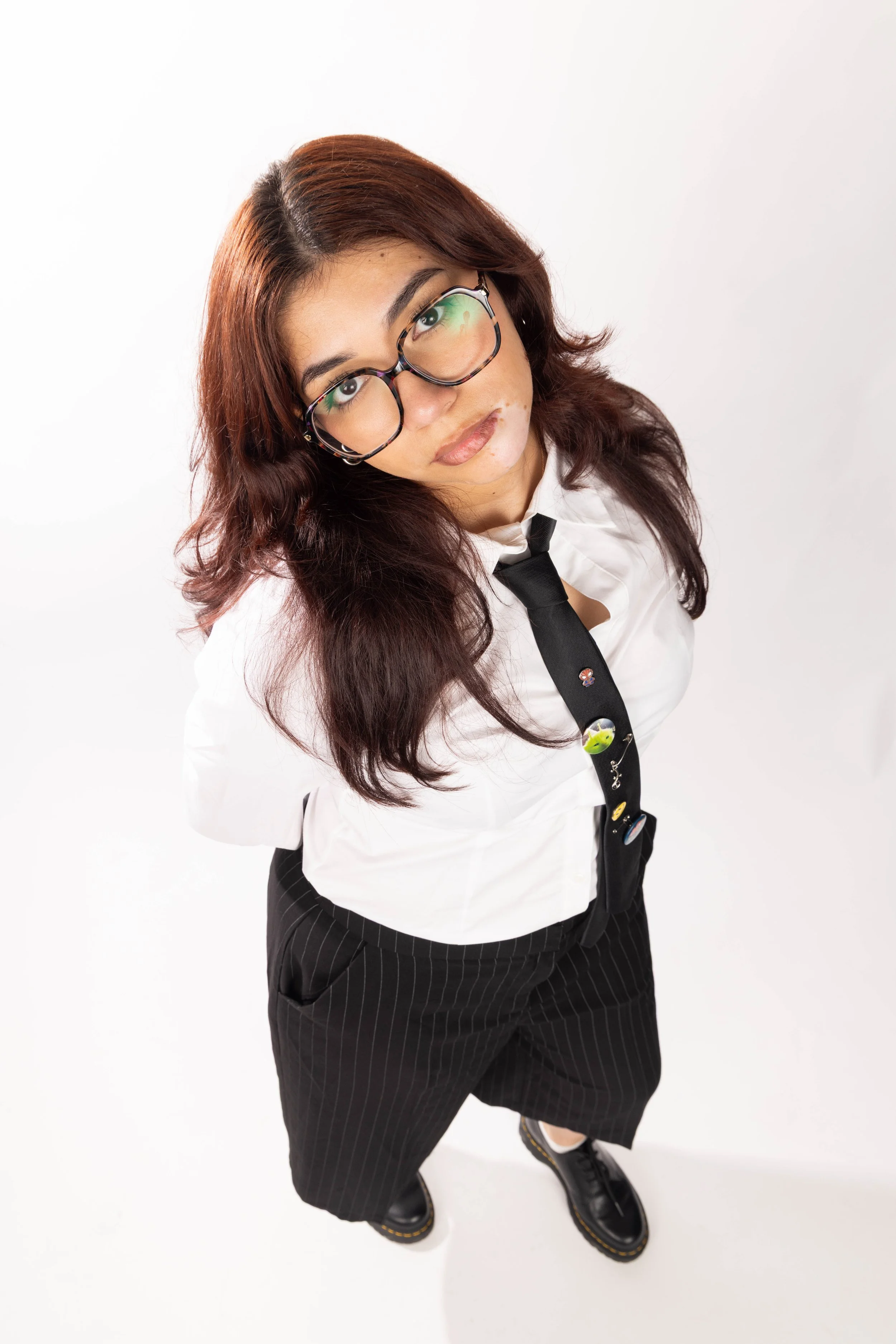 Woman with long reddish-brown hair, wearing large glasses, a white shirt with a black tie decorated with colorful pins, black pinstriped pants, and black shoes, standing against a plain white background.
