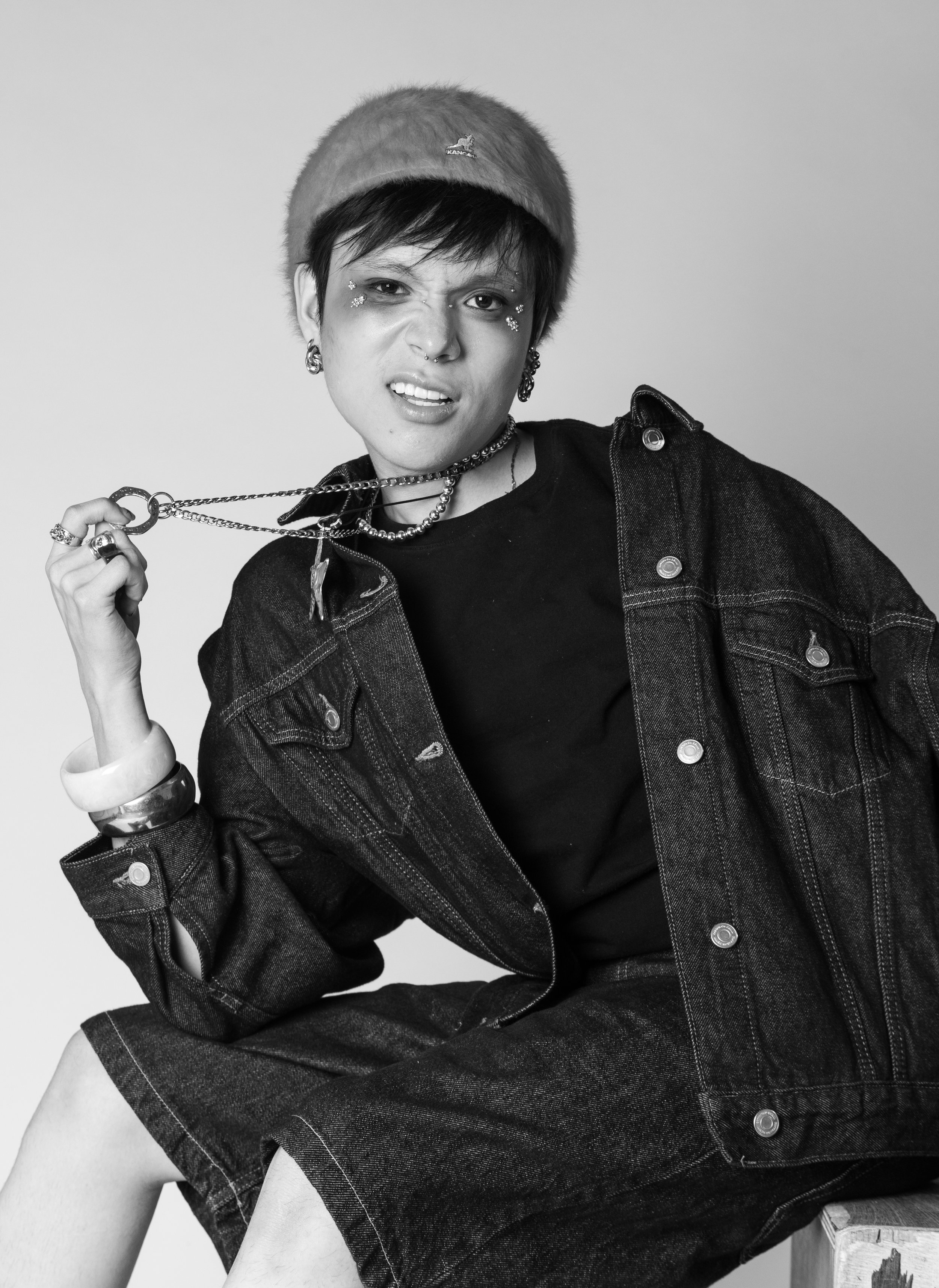 A person wearing a fluffy hat, denim jacket, and multiple jewelry pieces, making a playful facial expression.