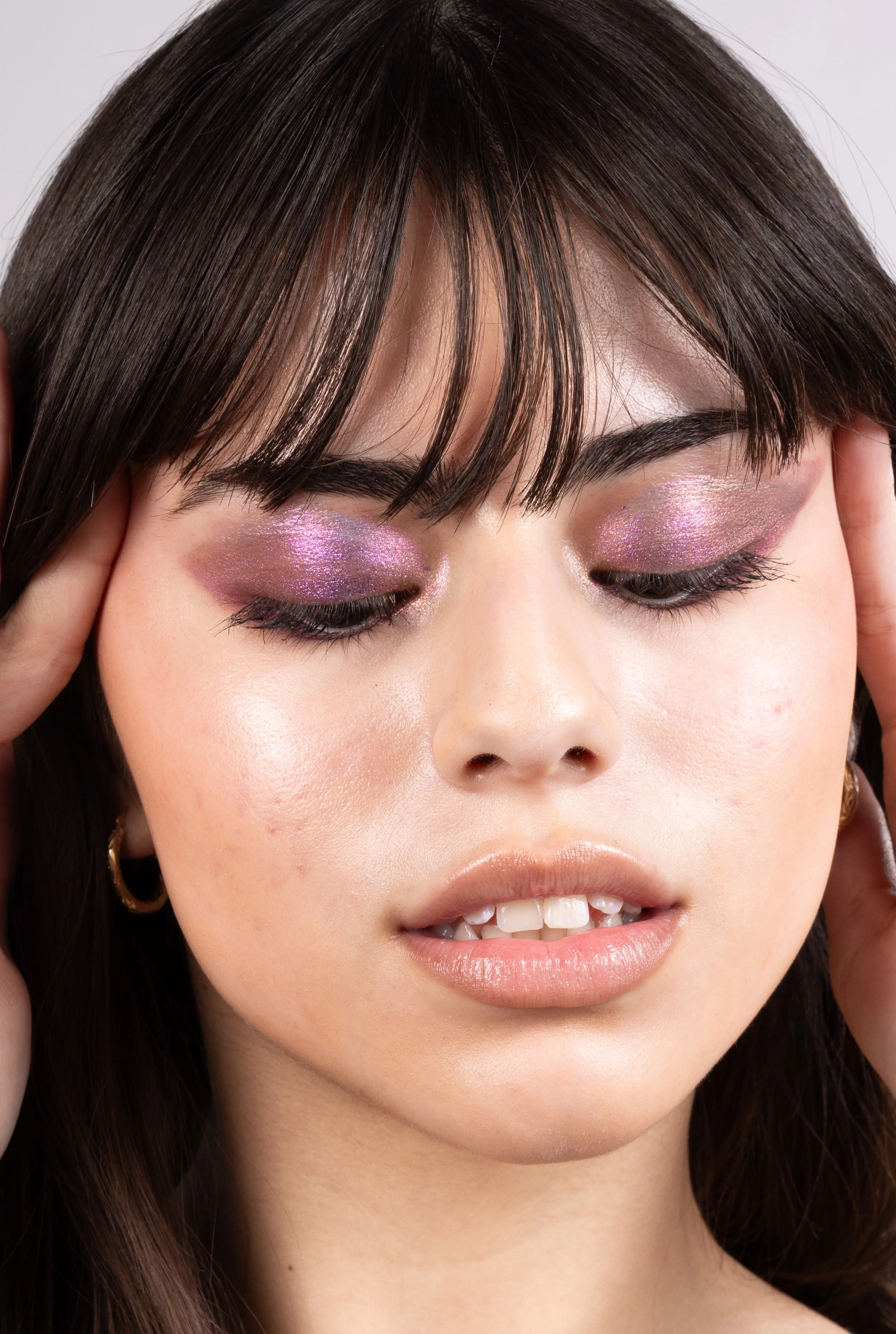 Close-up of a woman's face with closed eyes, wearing shiny pink and purple eyeshadow, with hands gently touching her temples.