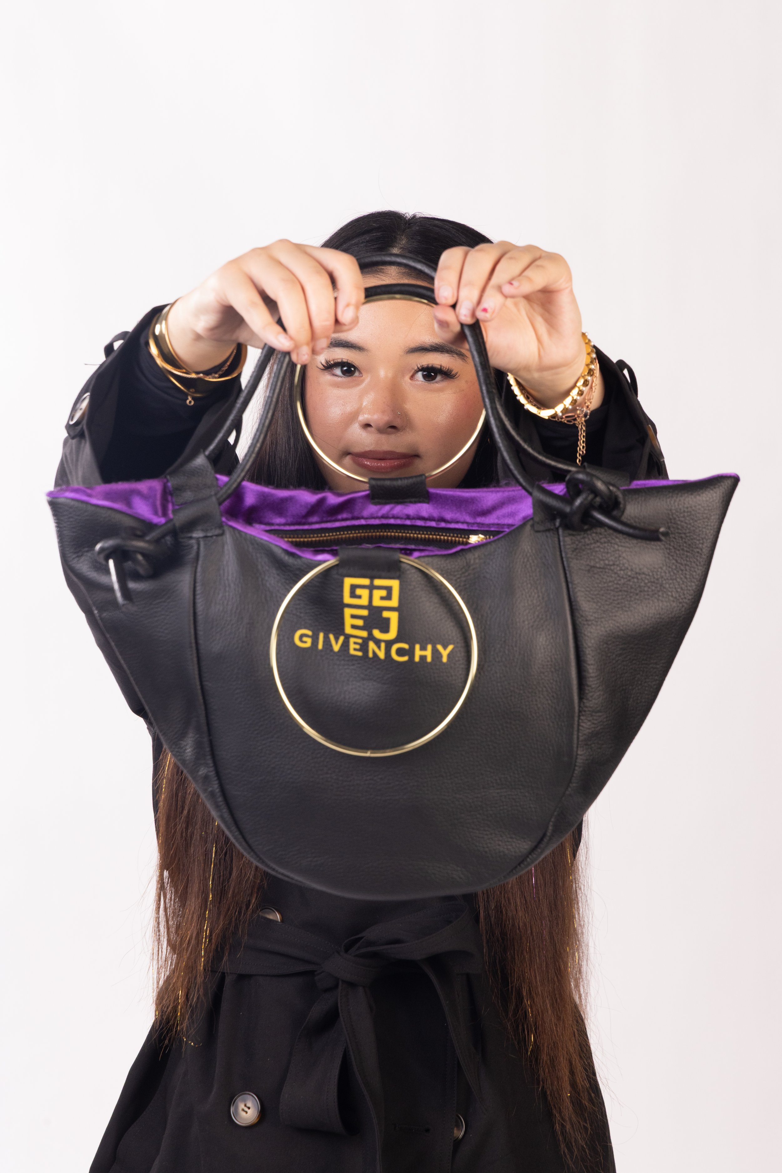 A woman holding a black Givenchy handbag with purple lining, looking directly at the camera, wearing gold jewelry, with long straight hair and a black outfit against a plain background.