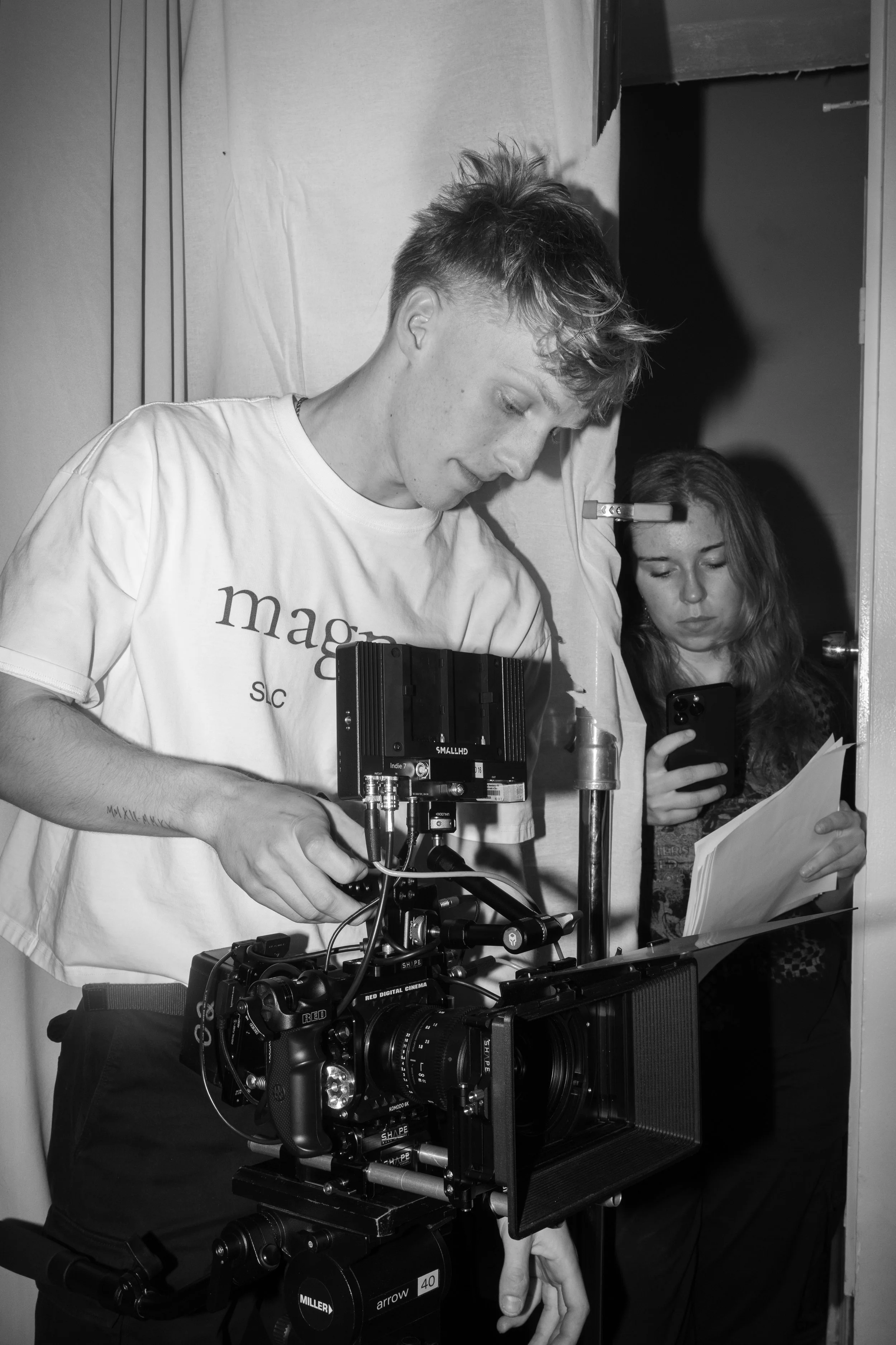 Two young people on film set, one adjusting camera, the other reading a script, black and white photo.