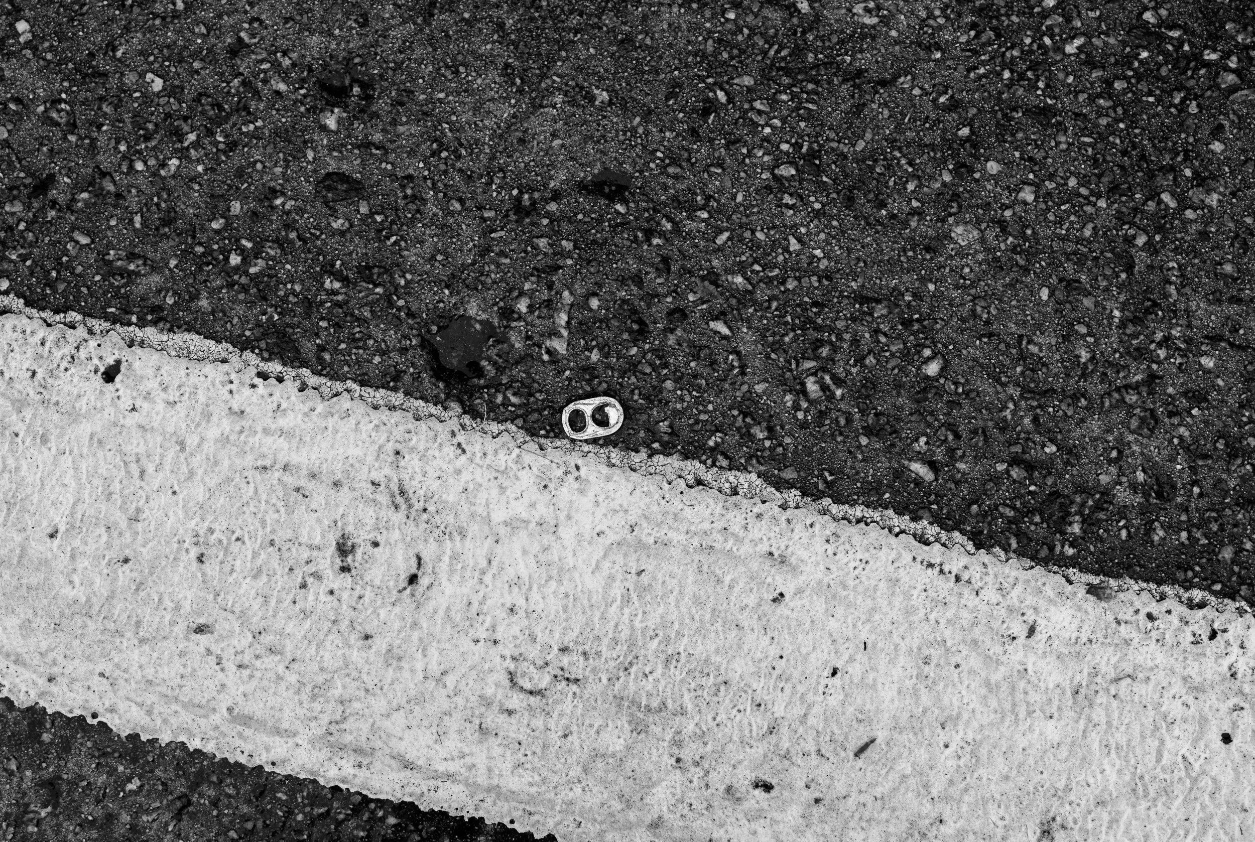 A close-up black and white photo of a road surface with a white painted line crossing over dark asphalt. Near the line, there is a small piece of metal, possibly part of a can or bottle, embedded in the road.