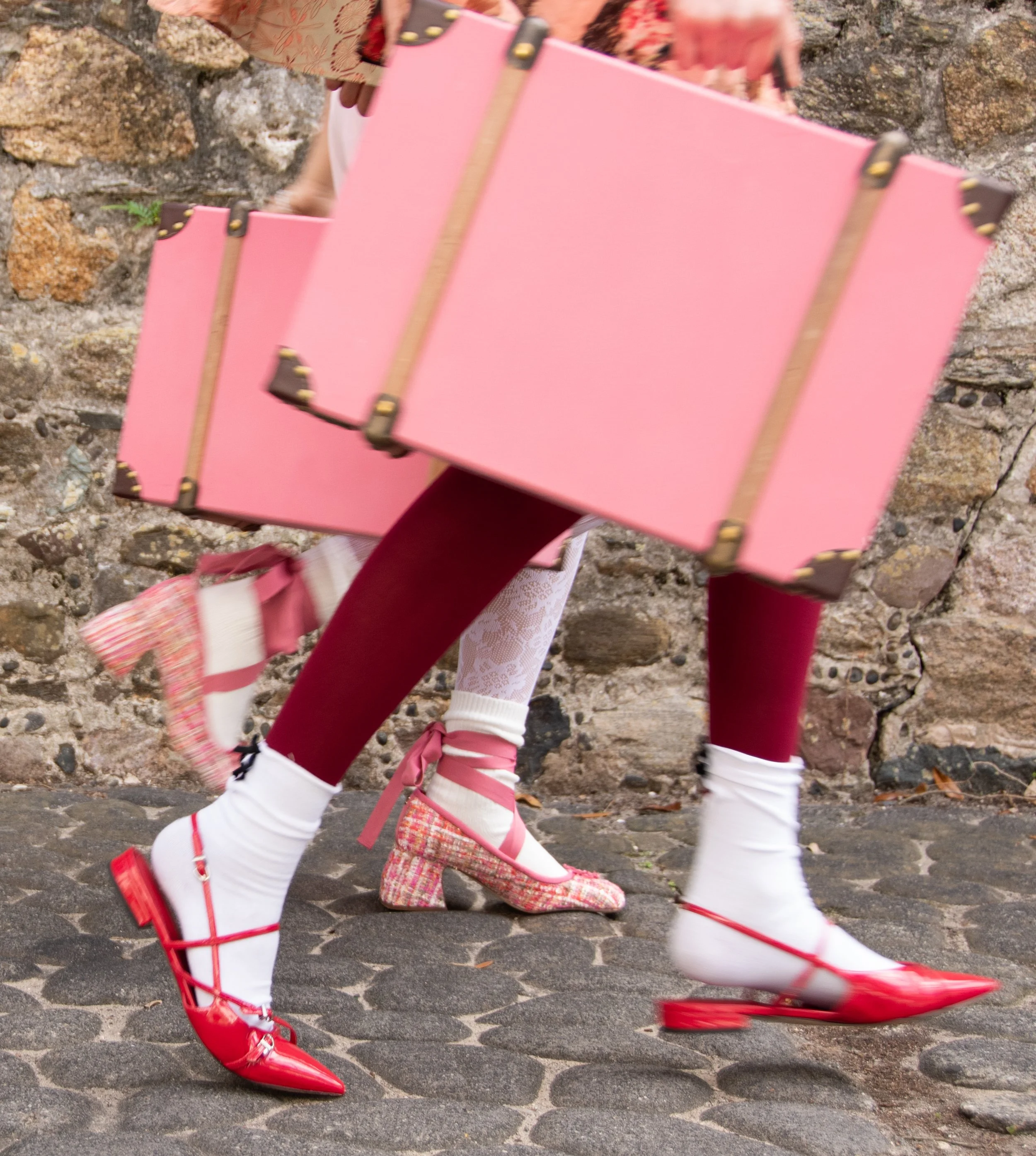 Close-up of a person carrying pink boxes, wearing red and white shoes, red tights, and white socks with pink and white lace patterns, walking on cobblestone street.