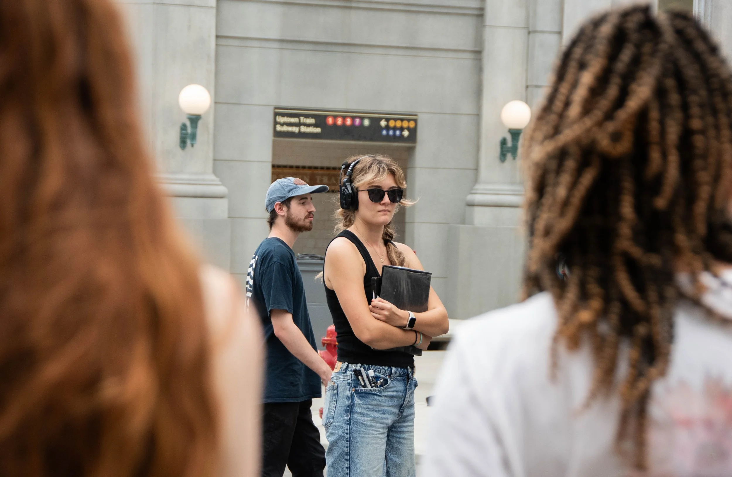 A woman with sunglasses, wearing a black tank top and jeans, standing with arms crossed in a subway station, surrounded by other people including a man with a baseball cap and a woman with braids.
