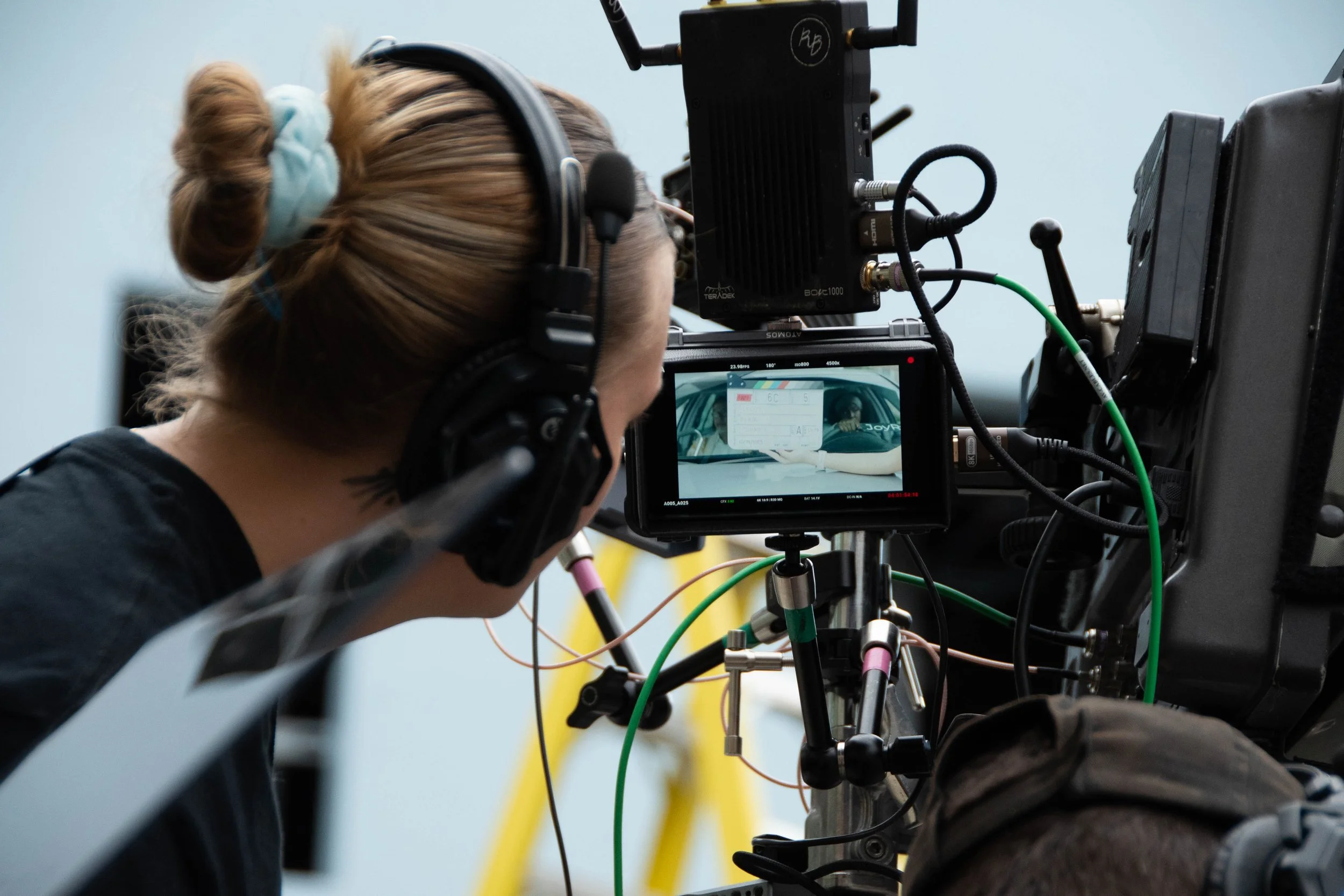 A woman wearing headphones and filming with a professional camera, focusing on a person holding a sign or card inside a vehicle.