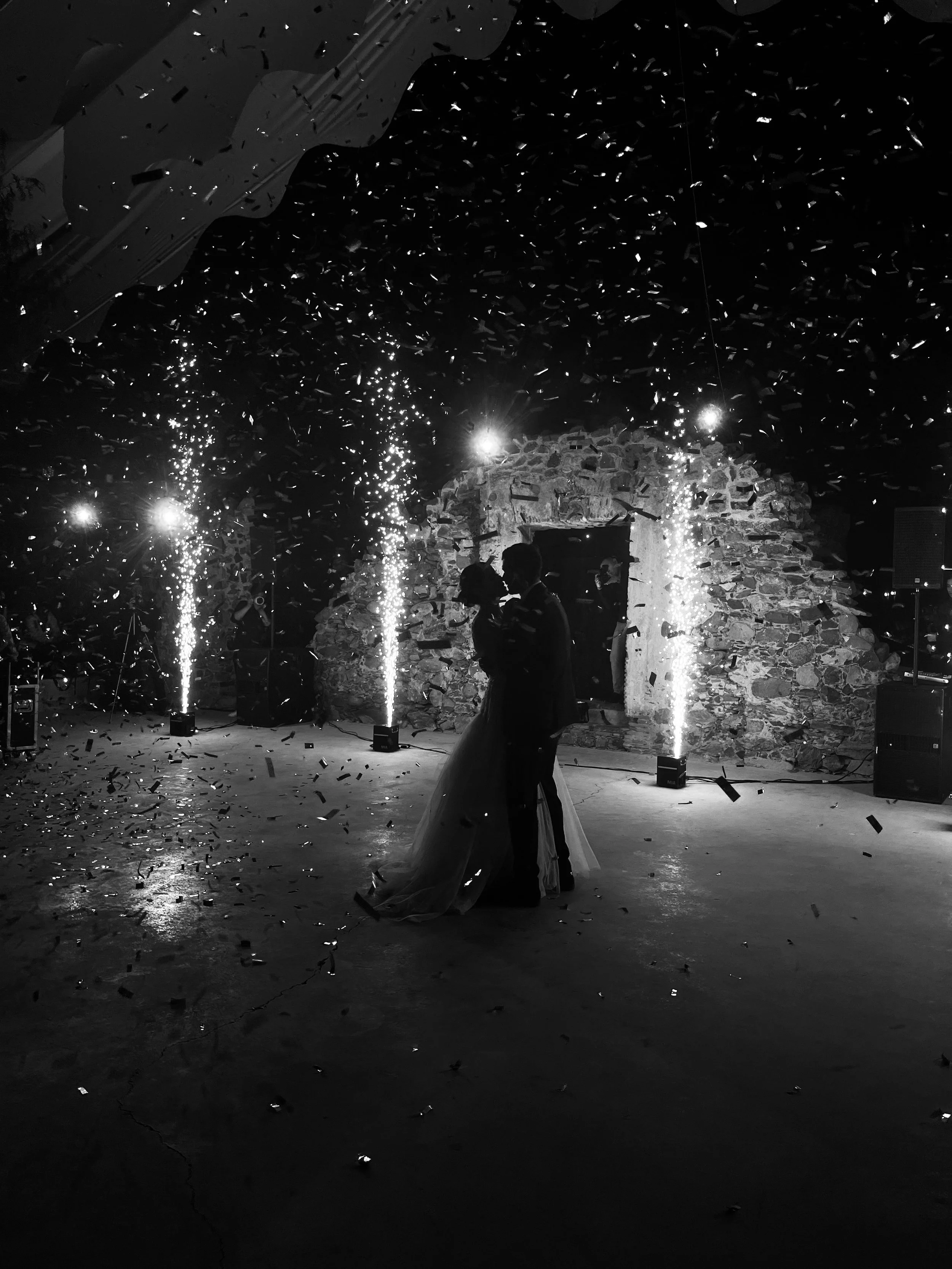 Silhouettes of a couple dancing at their wedding reception with fireworks and confetti in the background, stone wall and lighting creating a romantic atmosphere.