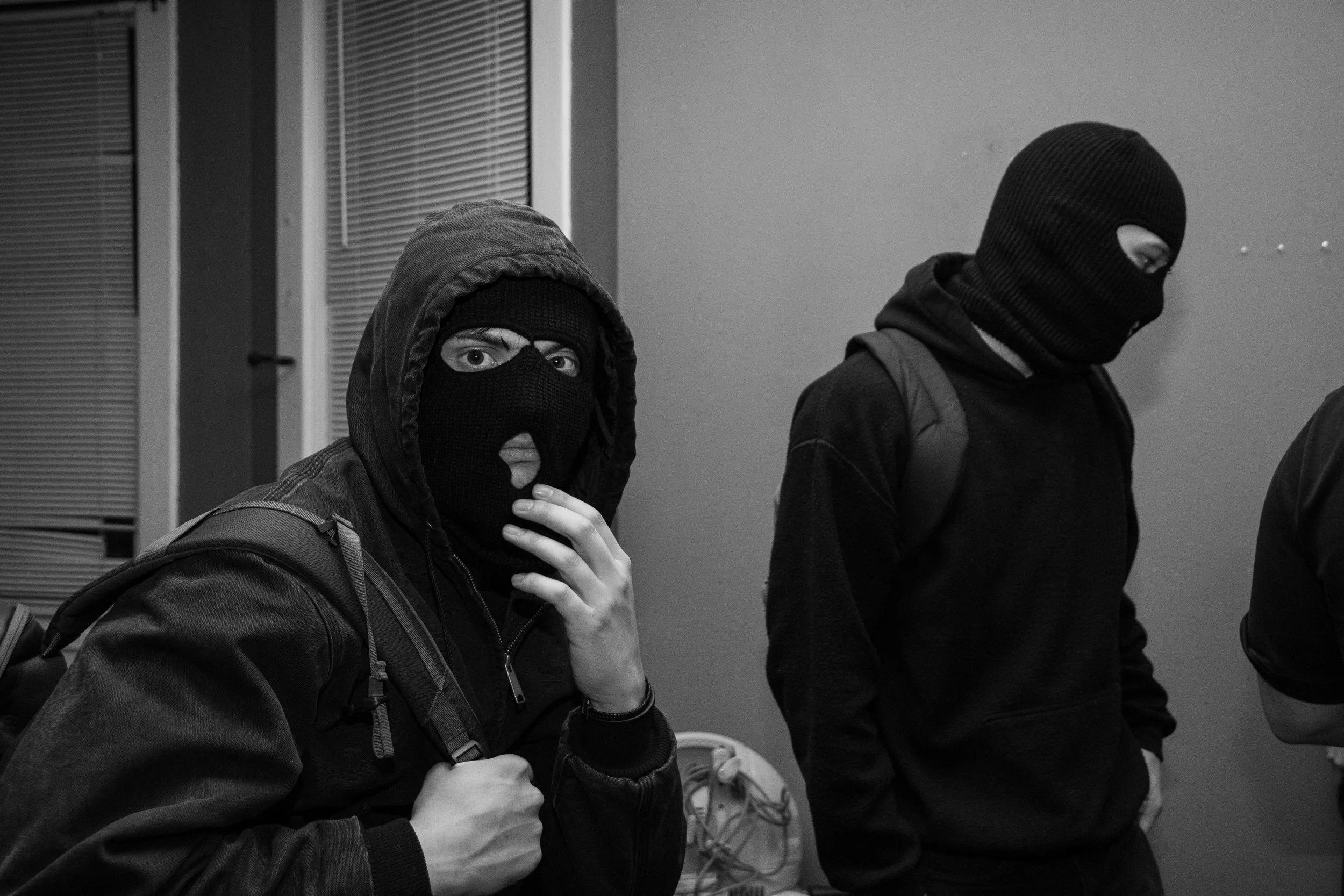 Two individuals wearing black ski masks and dark clothing indoors, one on the left adjusting his jacket and the other on the right looking down, with backpacks.