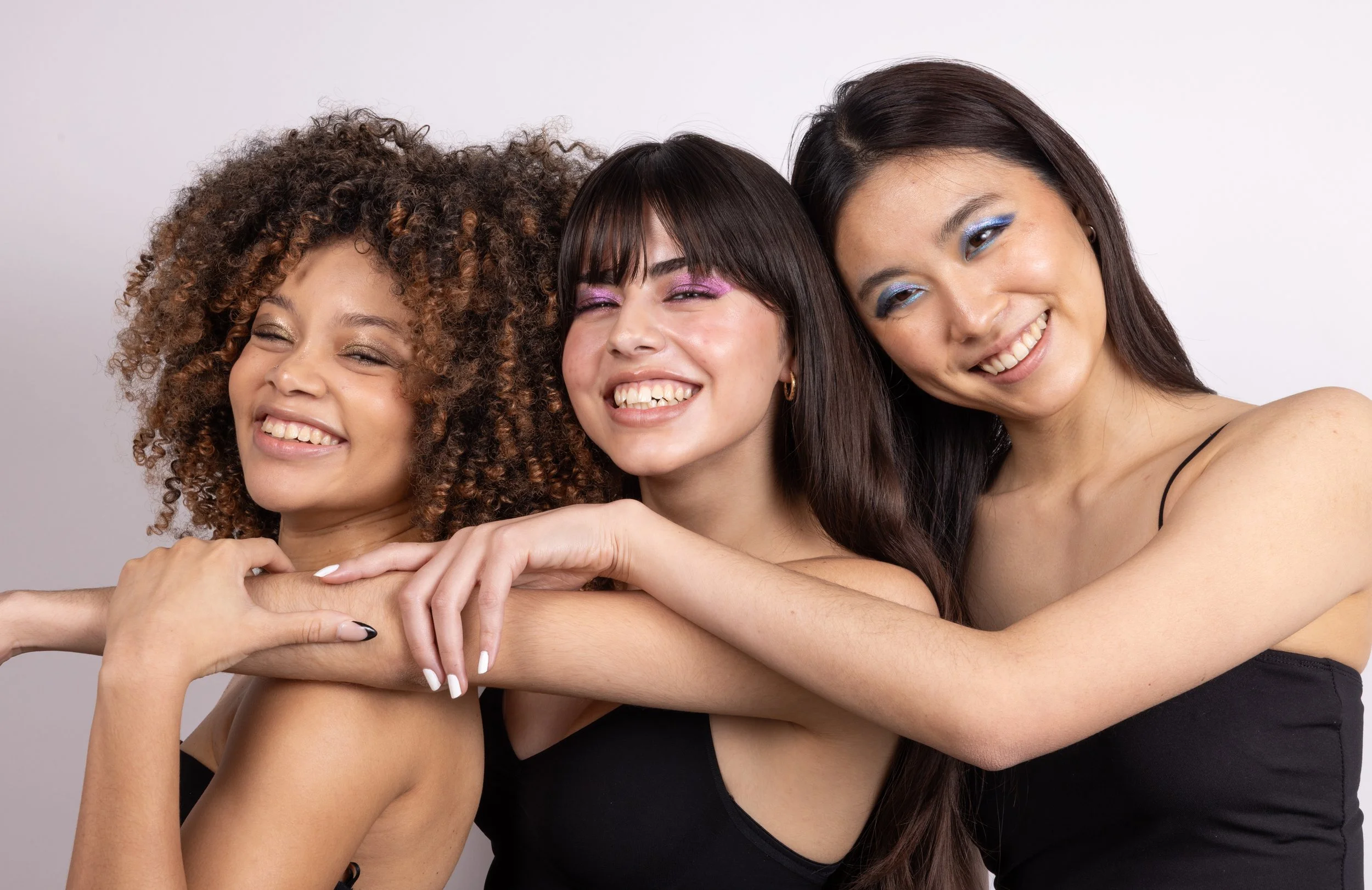 Three smiling women embracing each other in a close group shot against a plain background.