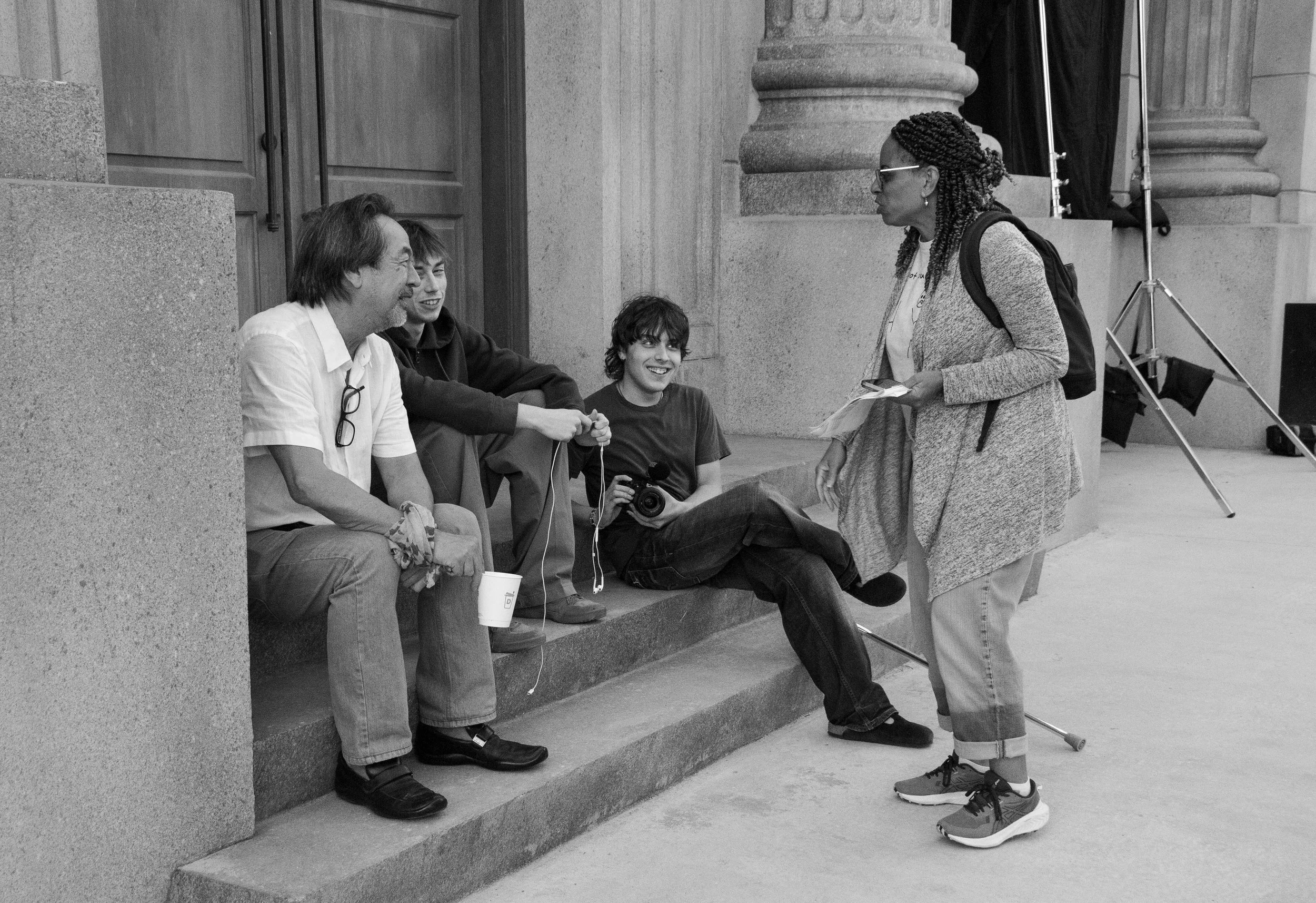 A woman with braided hair, glasses, and casual clothing talking to a group of four young men sitting on steps in front of a large stone building. The men are listening and smiling, one holding a camera and another with a cup. The woman is holding pap