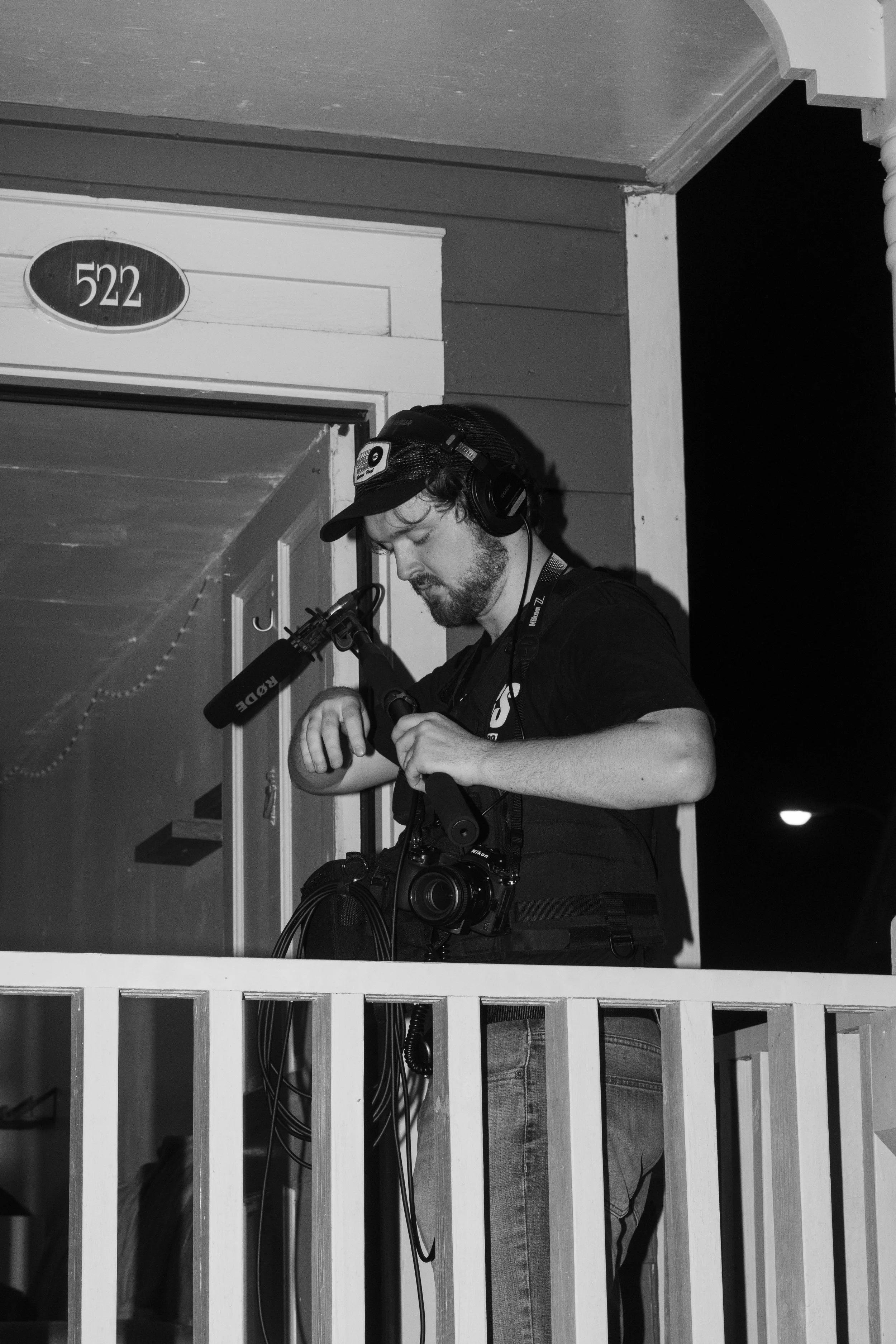 A man wearing a cap and headphones, holding a microphone, setting up or adjusting camera equipment on a balcony outside a house at night.