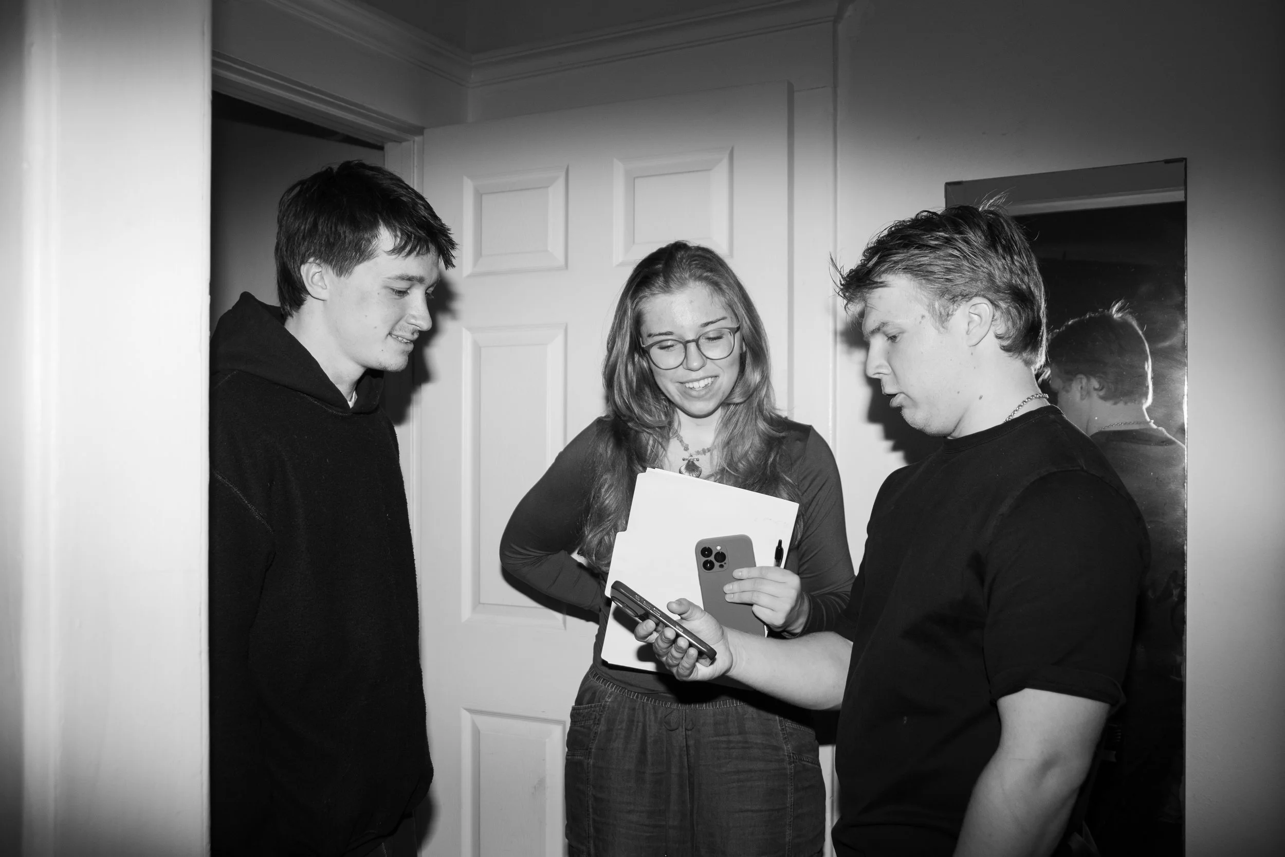 Three young people standing together indoors, looking at a smartphone held by one of them, with a mirror reflecting their image.