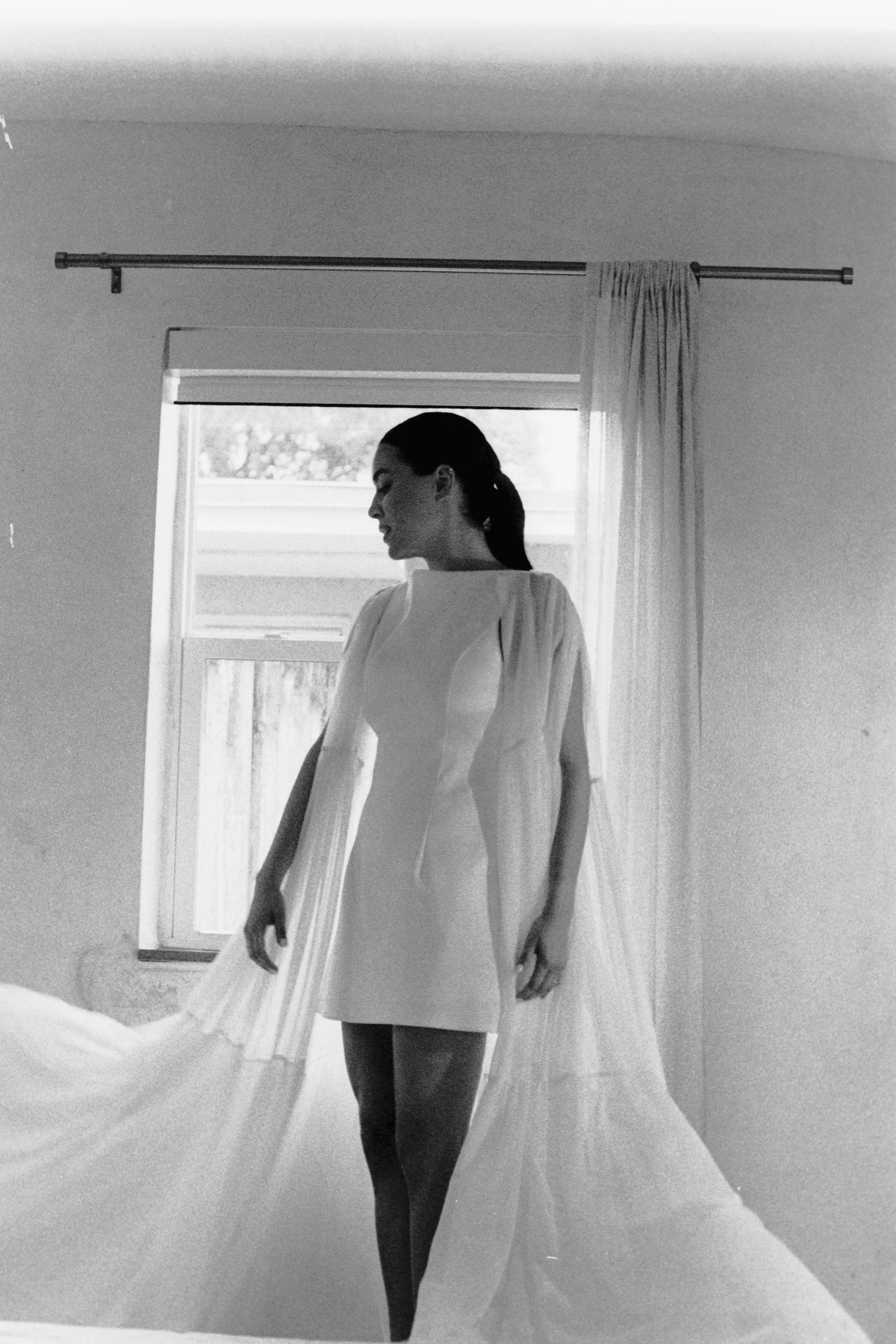 Black and white photo of a woman in a dress standing in front of a window with sheer curtains in a room.