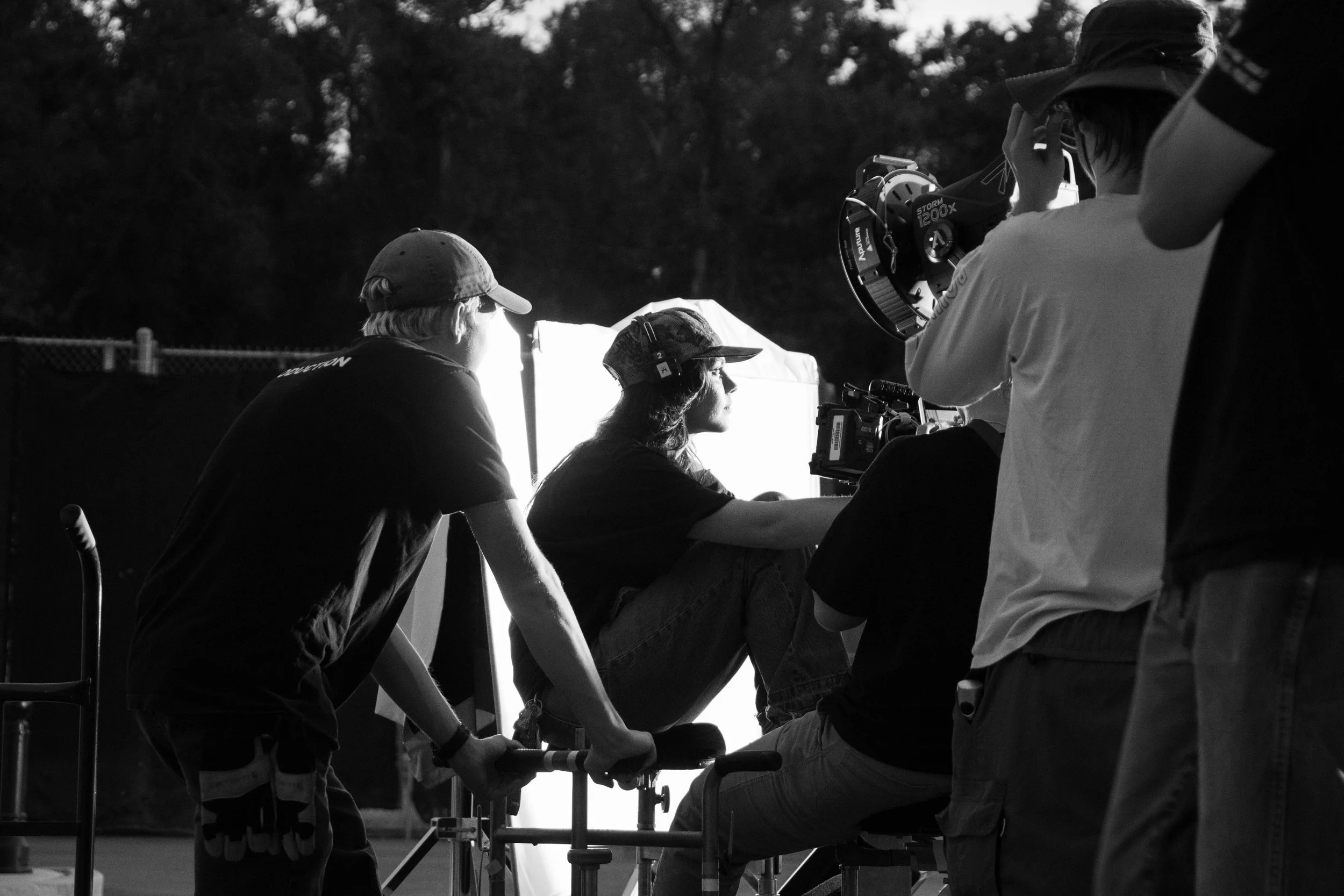 Filmmaking crew working outdoors on a film set with a person sitting on a dolly and multiple crew members around, filming with a camera.