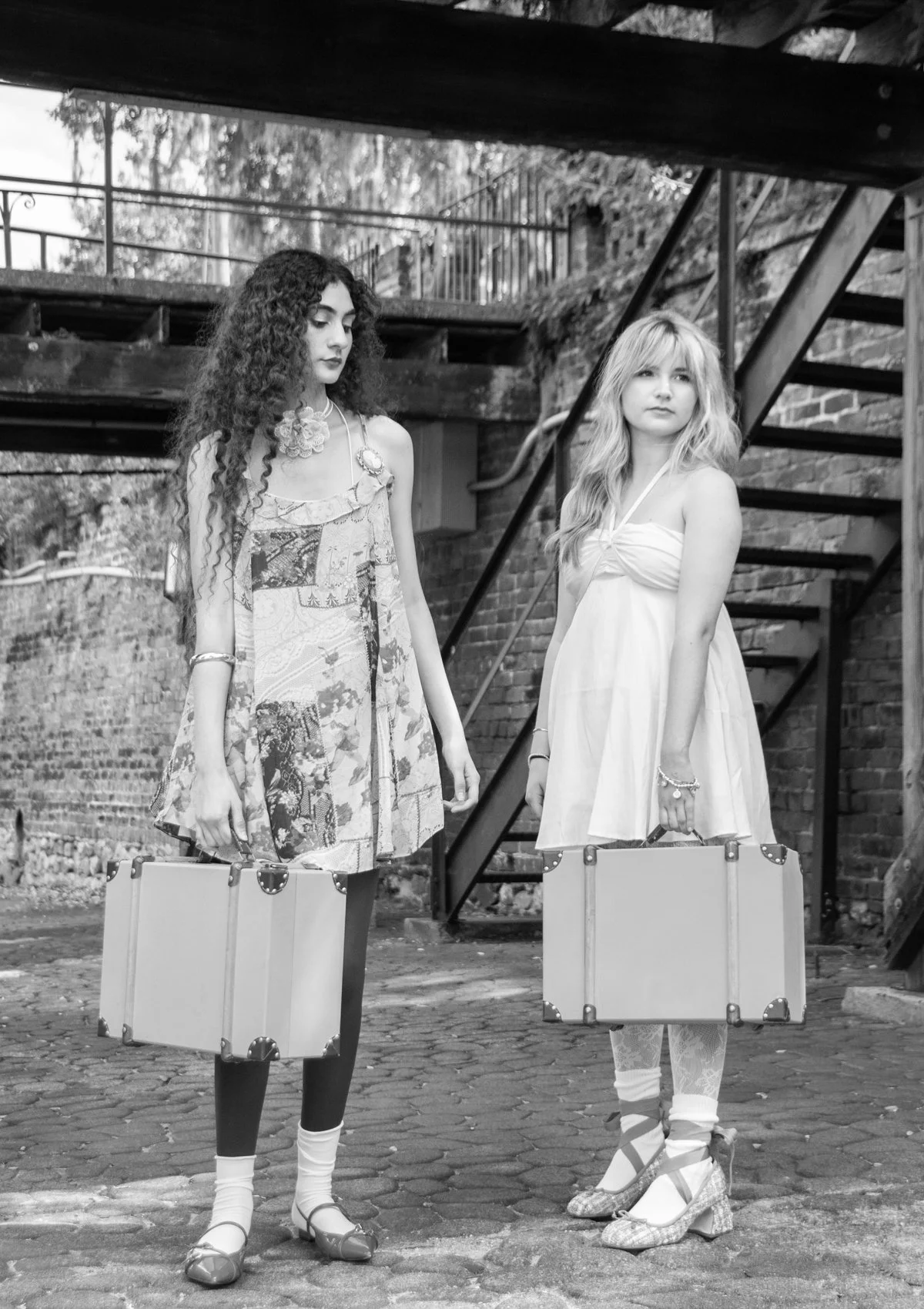 Two women standing on a cobblestone street, each holding a large suitcase, with a brick wall and a staircase in the background.