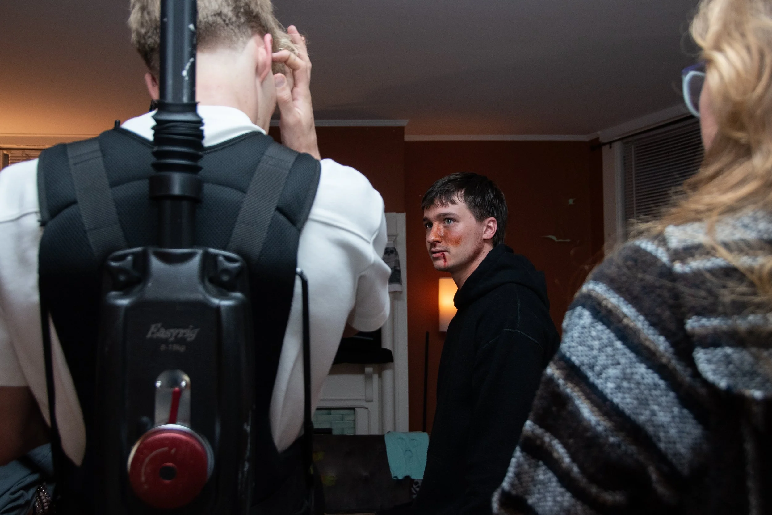 A young man with a bloody face and lips, looking at the camera, surrounded by two other people in a dimly lit indoor room.