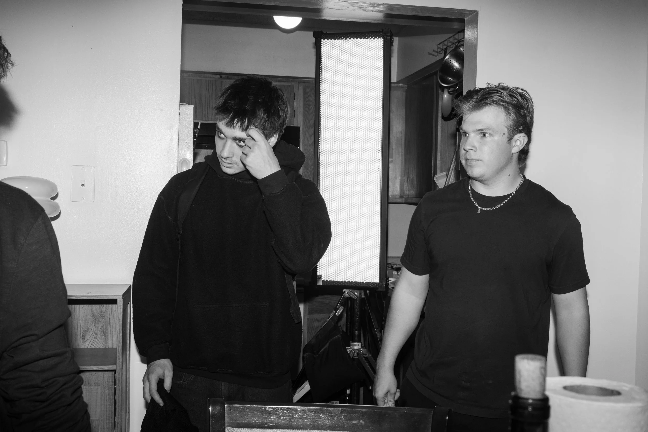 Two young men standing indoors, one adjusting his hair and the other looking to the side, with kitchen cabinets and a lighted screen in the background.