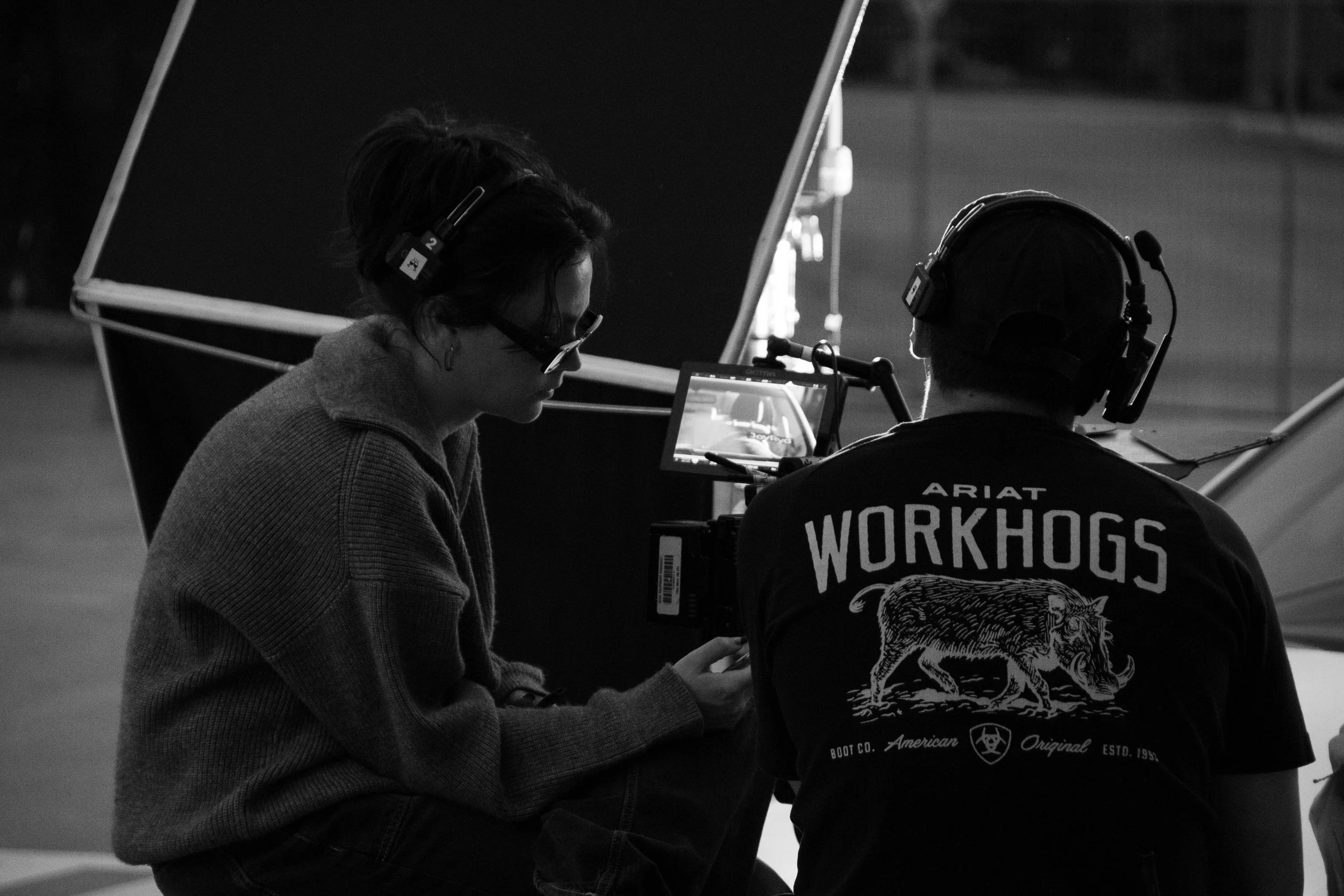 Two people wearing headsets on a film set, looking at a monitor, with a black and white background.